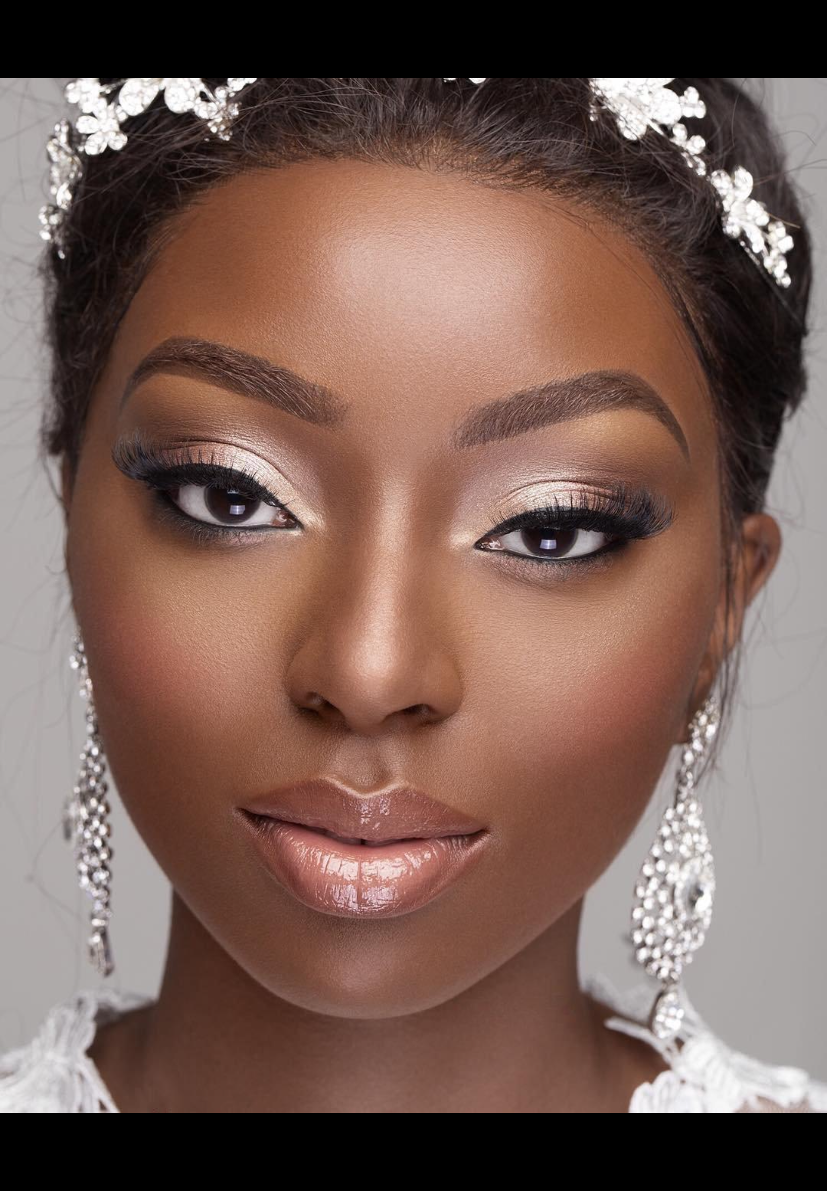 Close-up of a woman with makeup makeup, diamond earrings, white lace dress, and a jeweled headpiece, with a neutral gray background.