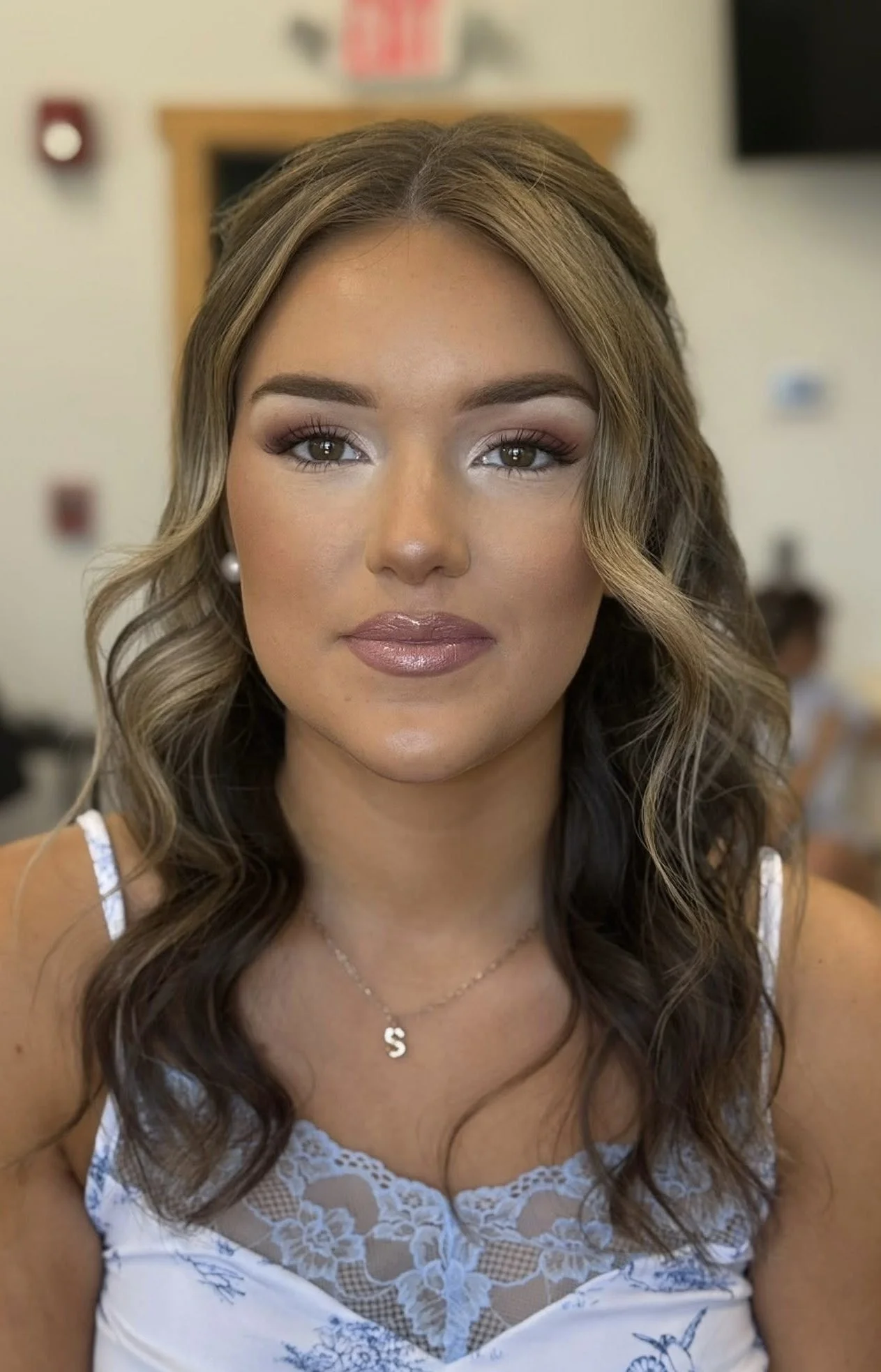 Close-up of a woman with light brown curly hair wearing a white lace top and a necklace with a letter 'S'. She has makeup on and is in an indoor setting.