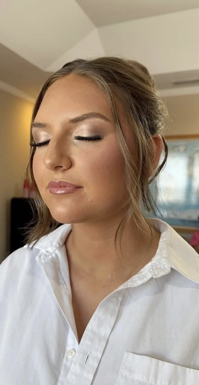 Close-up of a woman with makeup, eyes closed, wearing a white shirt, in an indoor setting with light-colored walls and window.