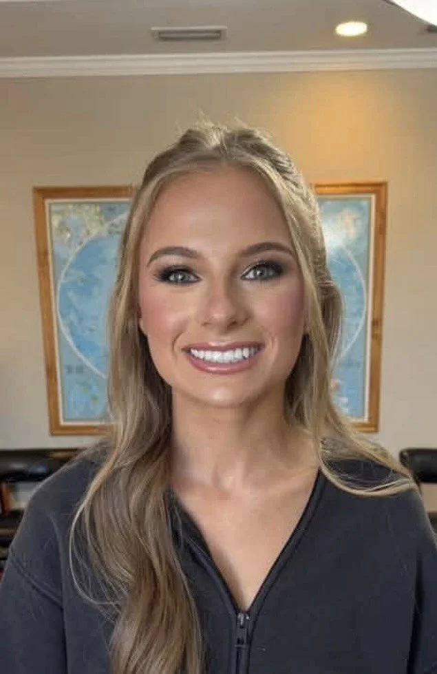 A smiling woman with long blonde hair and blue eyes, wearing a black zip-up top, standing in front of a world map in a room with beige walls and wood trim.