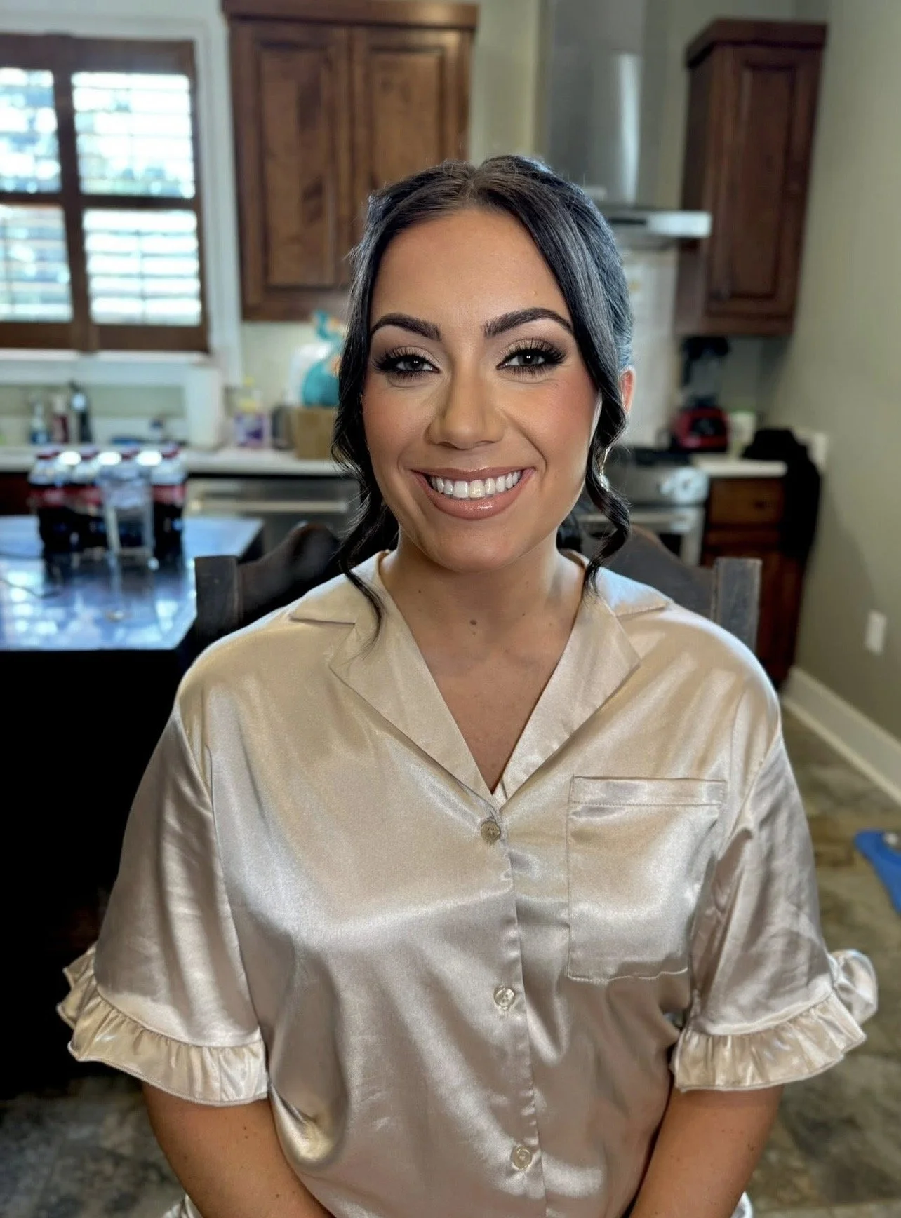 A woman with dark hair styled in loose curls, wearing a shiny beige satin pajama top with ruffled sleeves, smiling at the camera in a kitchen with wooden cabinets, a window with shutters, and various kitchen items in the background.