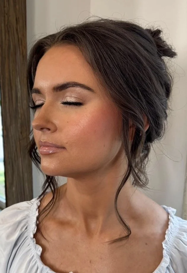 Close-up of a woman with dark hair styled in loose waves, closed eyes, natural makeup, and a white ruffled top.