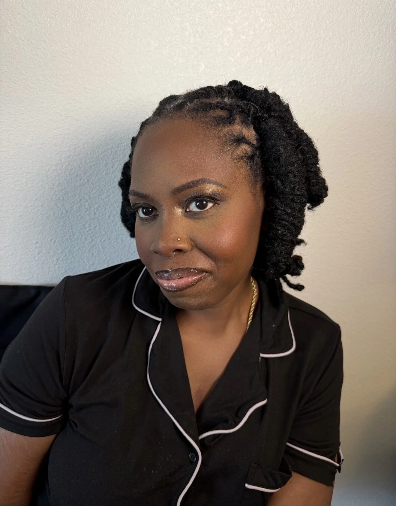 A woman with dark skin and braided hair styled into twists, wearing makeup with gold eyeshadow, and dressed in a black shirt with white piping confers a composed expression as she looks slightly to the left, seated against a plain off-white wall.