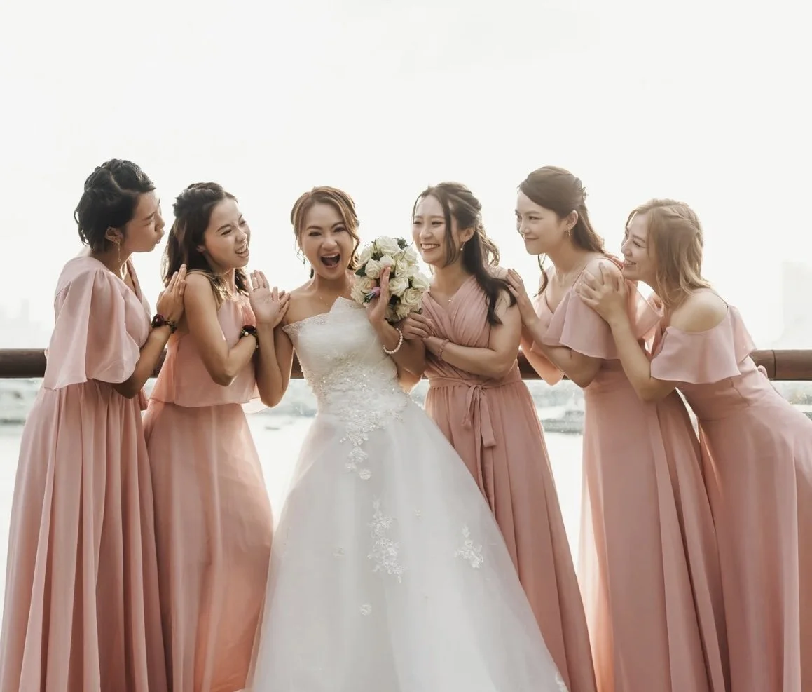 A bride in a white wedding gown holding a bouquet of white and pink flowers, surrounded by five bridesmaids in matching pink dresses, all smiling and happy outdoors with a city skyline and harbor in the background.