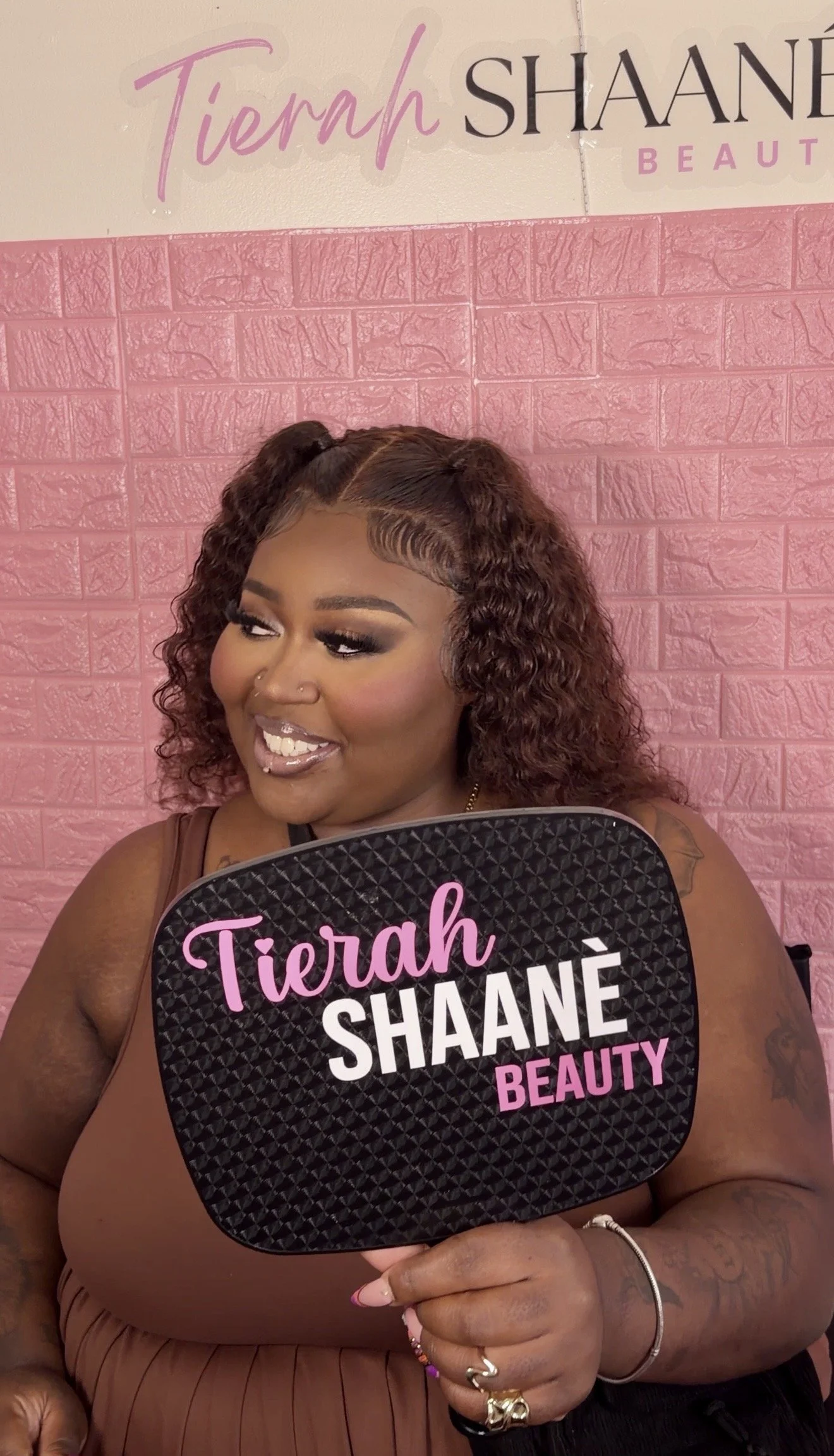 A woman with curly hair and makeup holds a sign that reads "Tierah Shaanè Beauty." She is smiling and standing in front of a pink brick wall with part of a sign visible above her head.