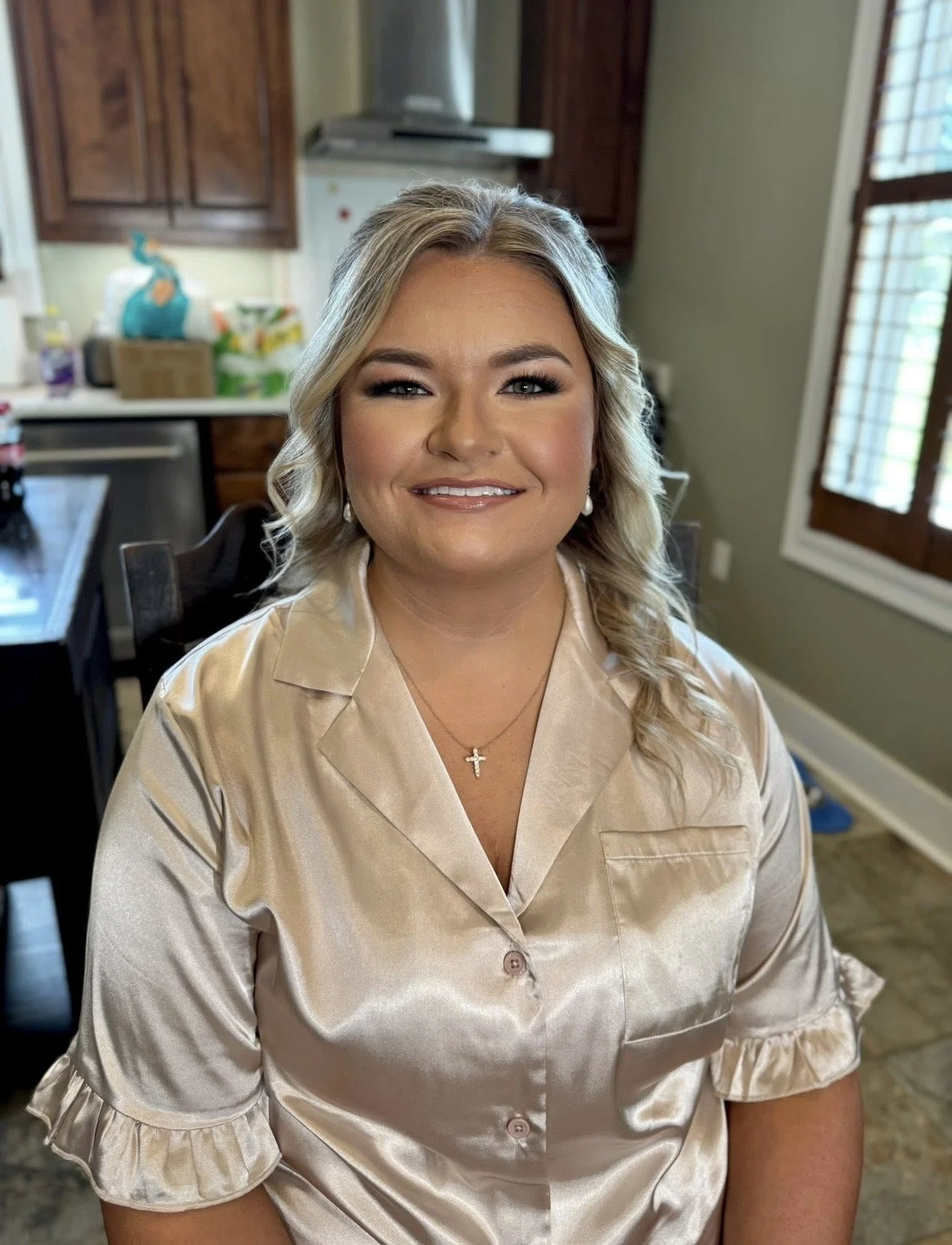 A woman with blonde, curly hair wearing a satin beige blouse and a necklace with a cross, smiling in a kitchen.