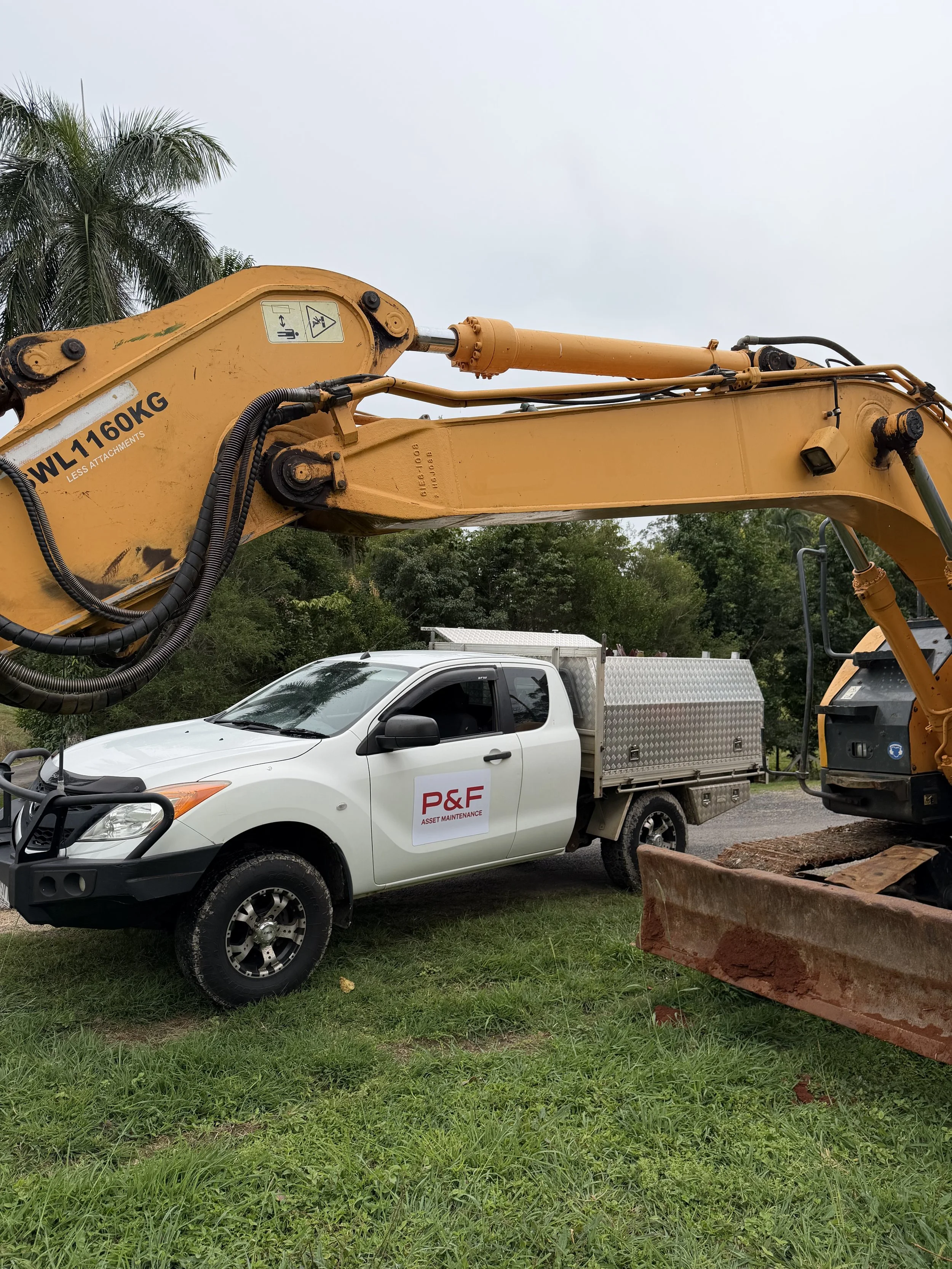 A white work truck with a yellow backhoe loader attachment parked on a grassy area near a dirt road. The vehicle has a P&F Asset Maintenance logo on it's door. In the background, there are cows on a hillside and trees, local Australian farmer's land.