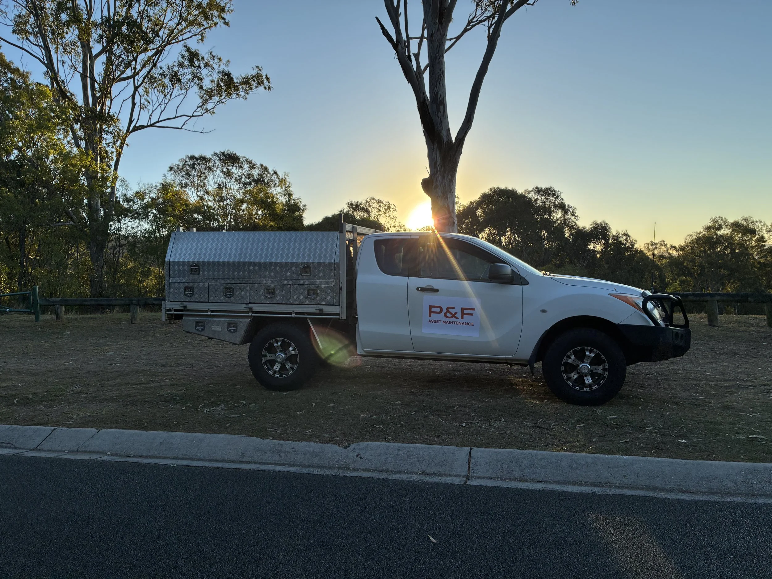 A white work truck with a silver toolbox in the bed, with P&F Asset Maintenance logo on vehicle's doors, parked on a curbside street during sunset, with trees and a clear sky in the background.