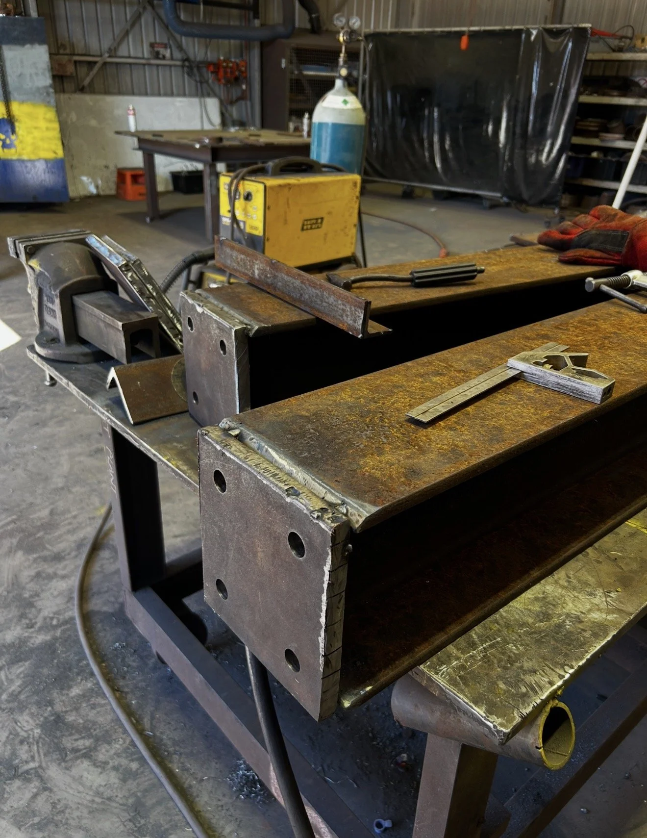 A workshop with a metal worktable, a yellow welding machine, a welding helmet, gloves, a measuring square, and a metal piece on the table, with various tools and equipment in the background.