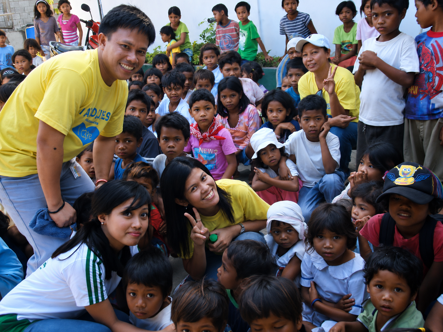 Group of children and three adults, some smiling and making peace signs, gathered outdoors for a community or school event, with some children sitting and others standing in the background.