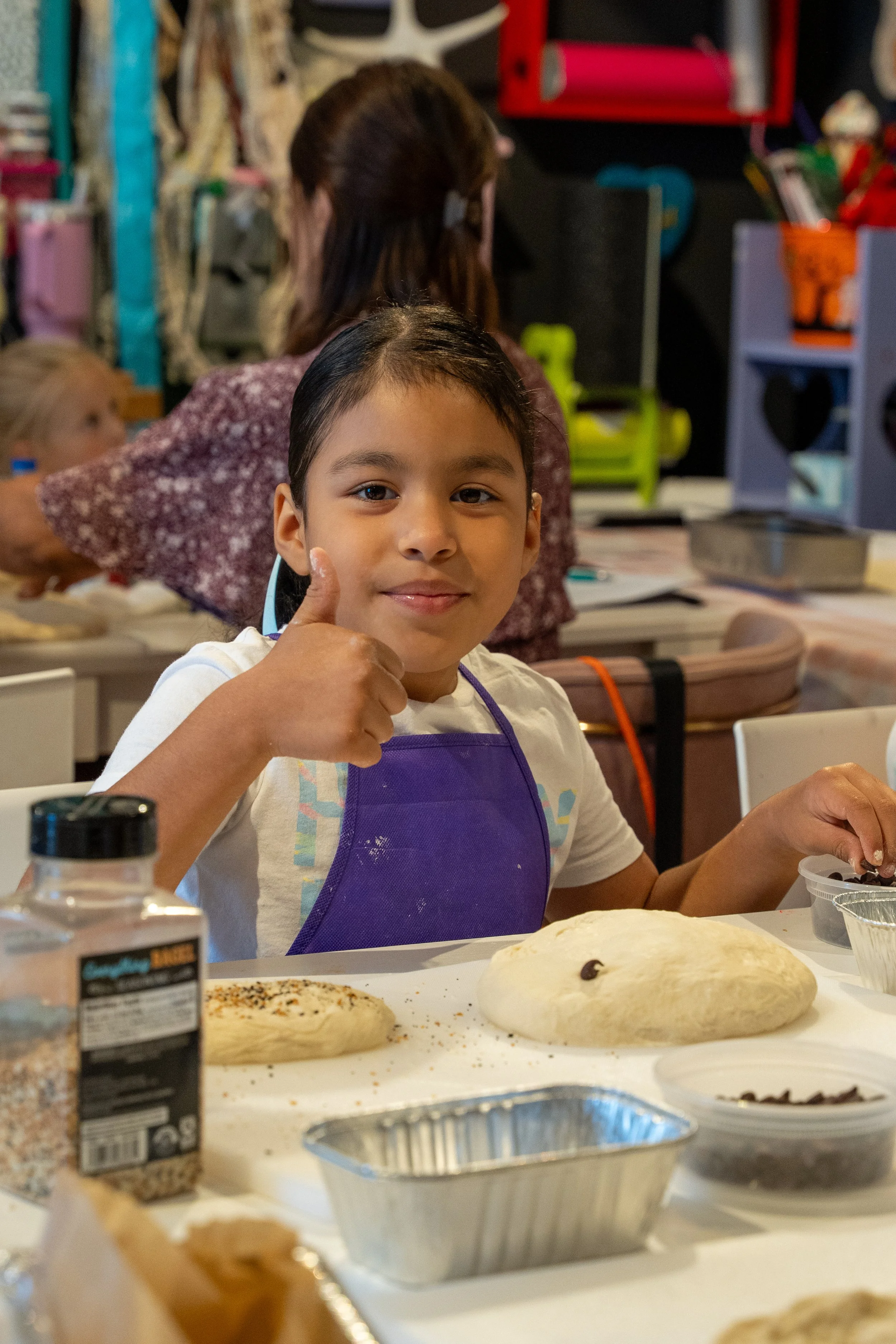 A young girl in a purple apron gives a thumbs-up during a sourdough class.