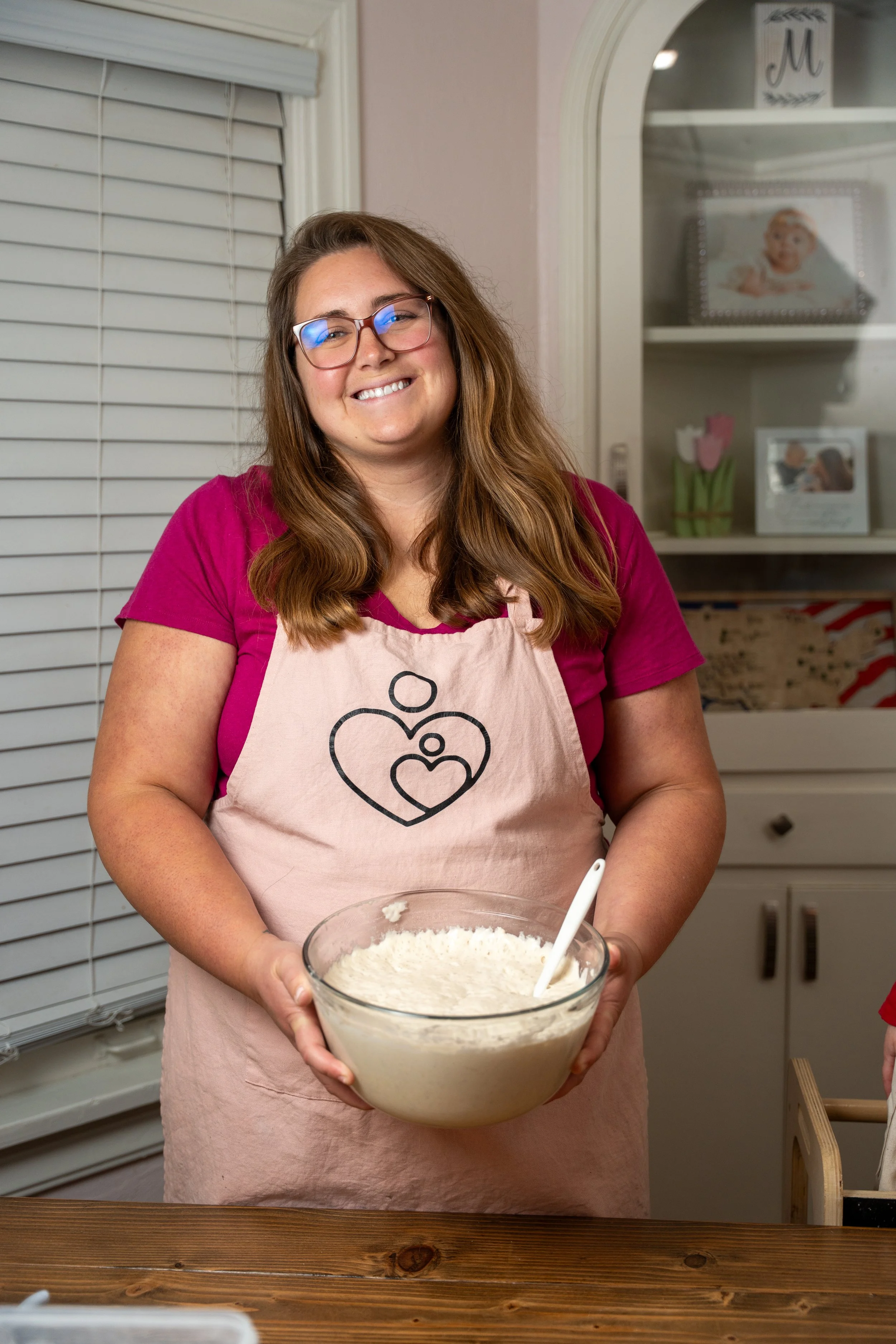 A woman holds a bowl of sourdough dough with a spatula in a kitchen.