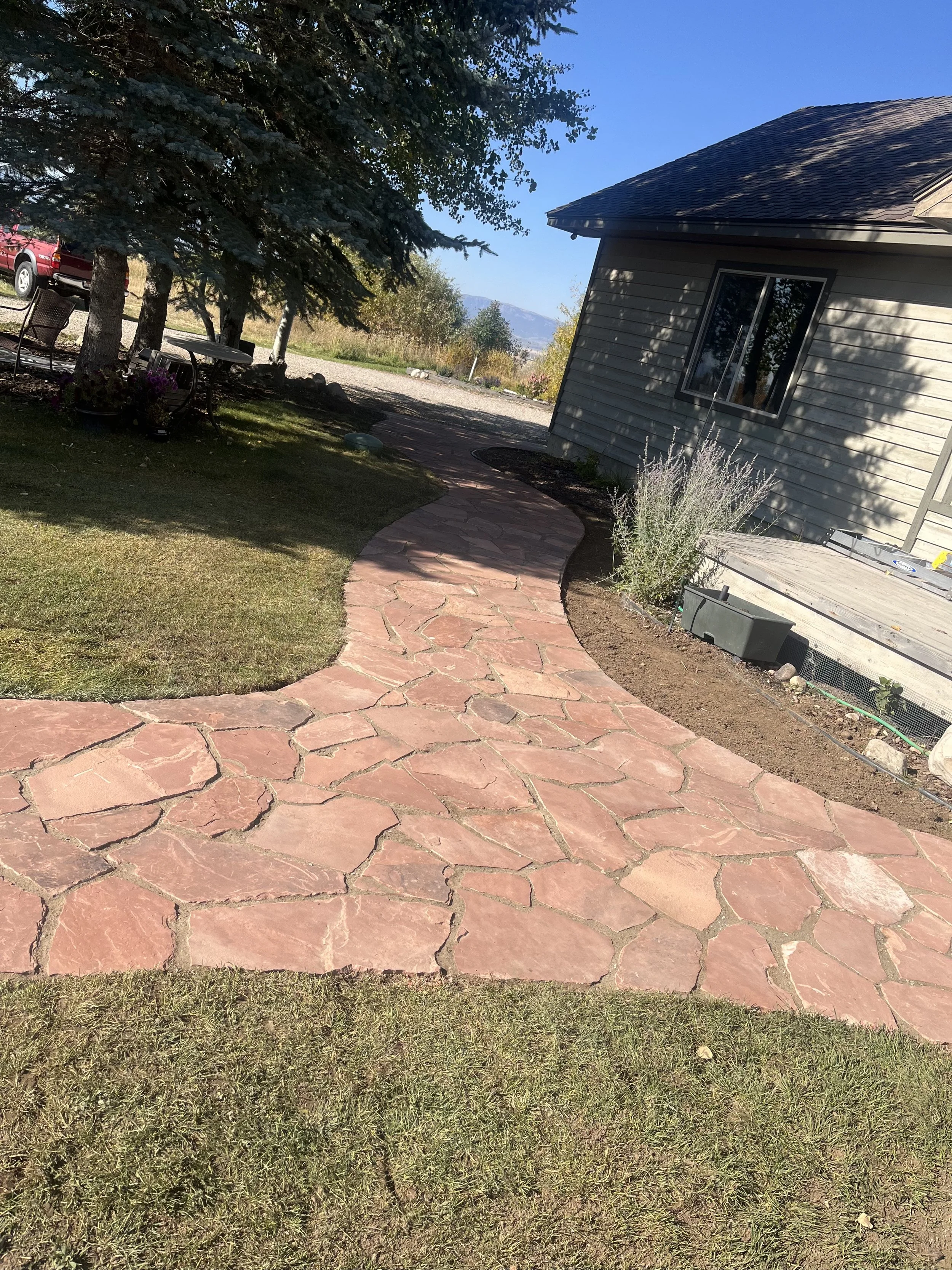 A curved red stone pathway running through a backyard with grass, trees, and a house on the right. A parked red truck and outdoor chairs are visible on the left. Mountains are in the background under a clear blue sky.