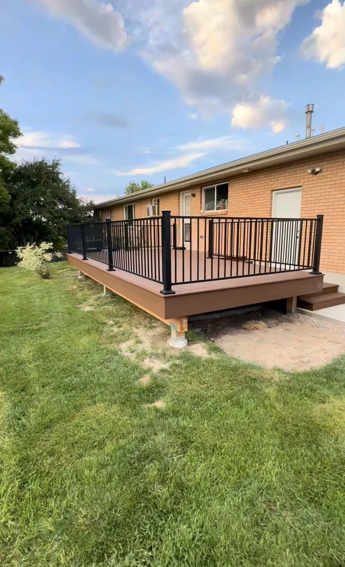 A newly built wooden deck with black metal railings extends from a brick house, surrounded by green grass, under a partly cloudy sky.