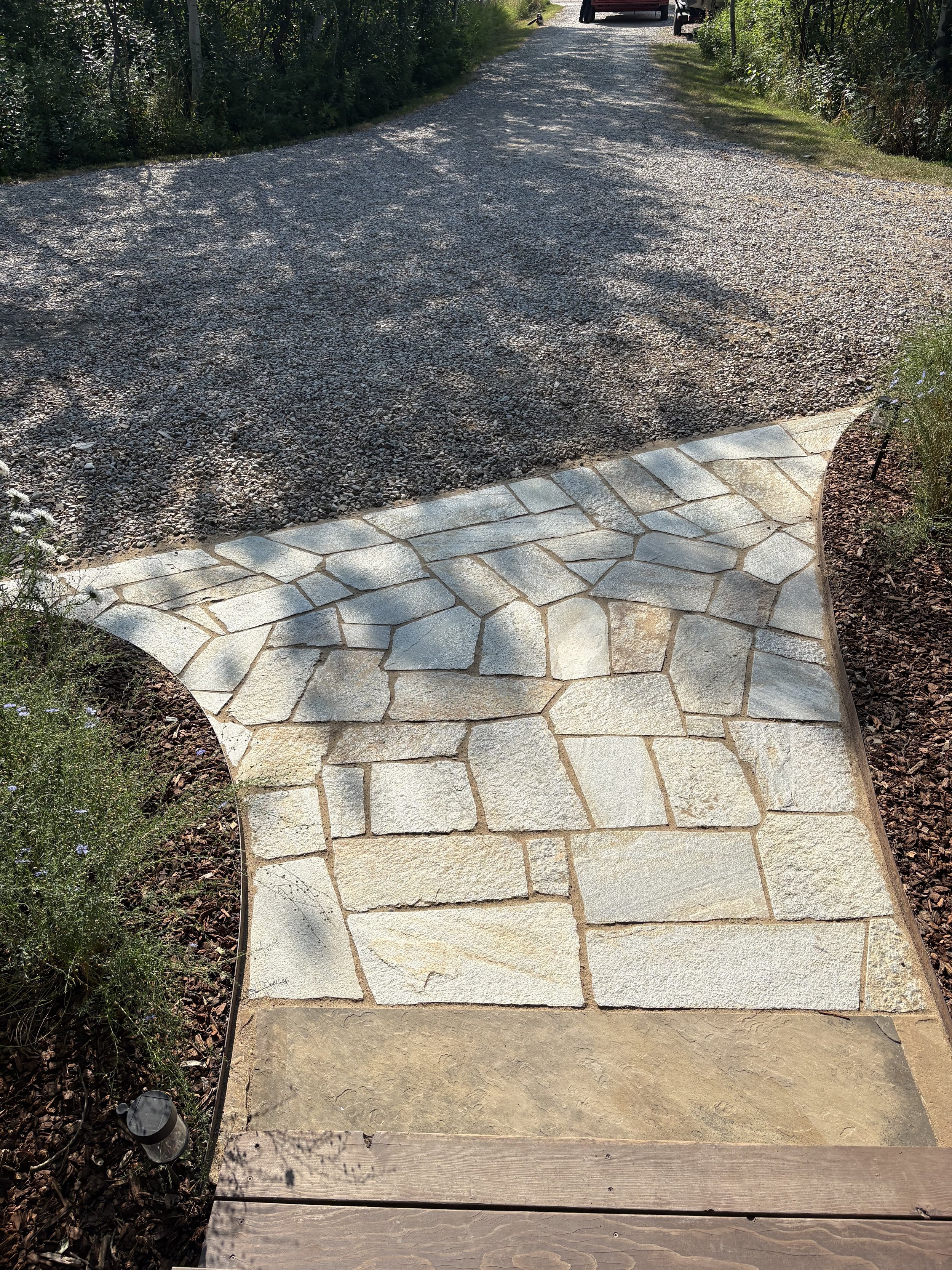 A pathway with a transition from flagstone to pavement, surrounded by greenery and bushes.