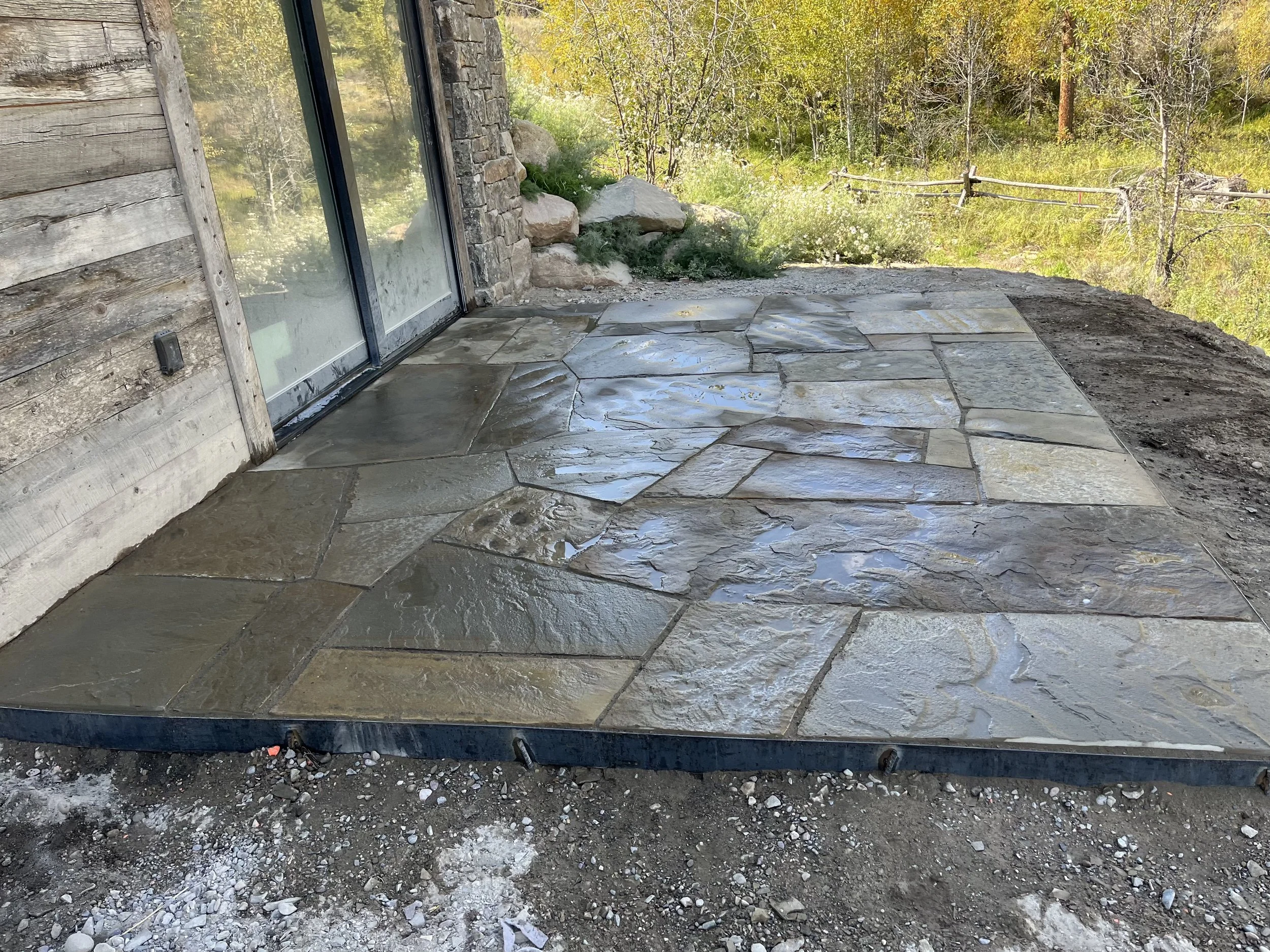 A stone patio area with a glass sliding door, next to a rustic wooden exterior wall, and surrounded by a natural outdoor landscape with trees and greenery.