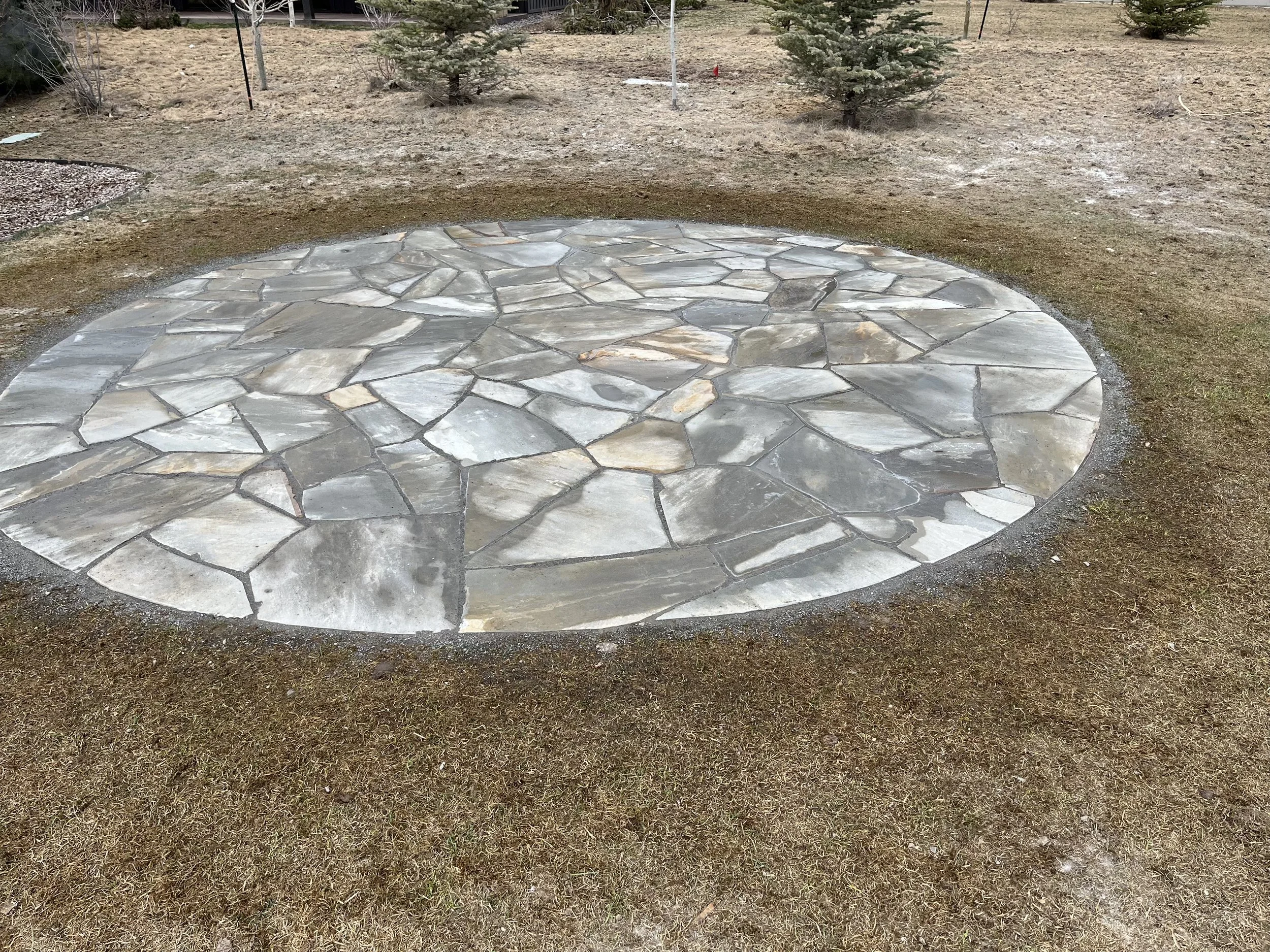 A circular area of flagstone pavers surrounded by bare dirt and sparse grass, with small evergreen trees in the background.