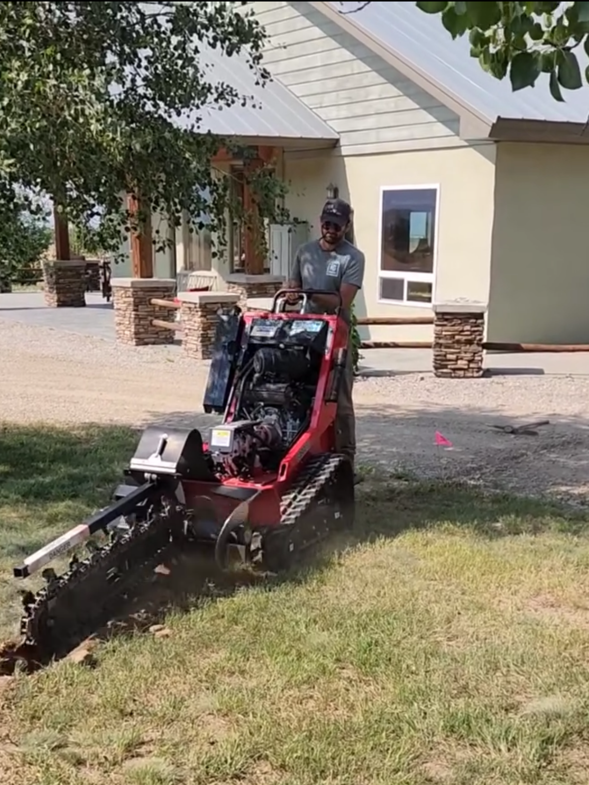 A man operating a roller compactor on a grassy yard in front of a house with stone pillars and a beige exterior.