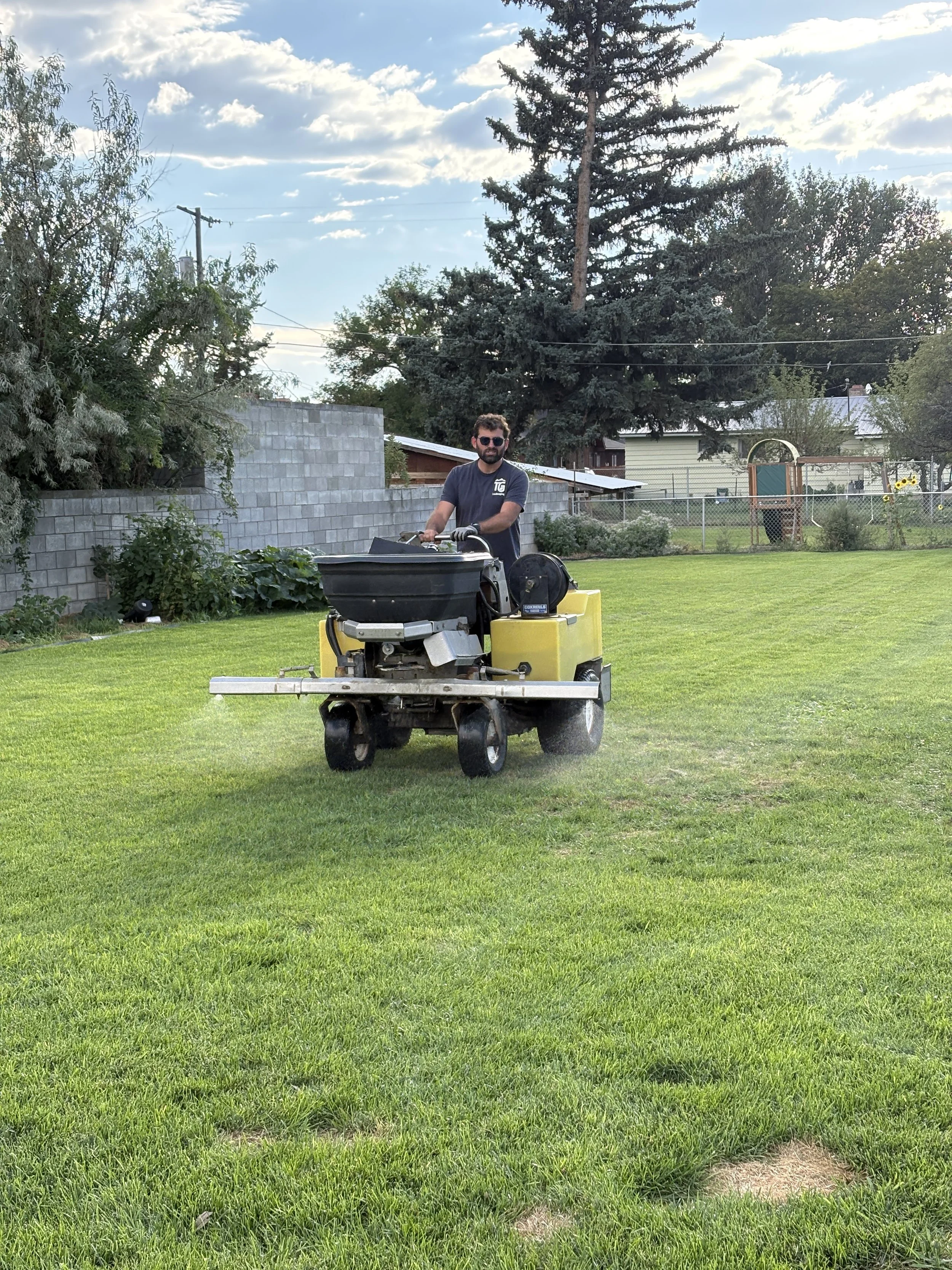 A man operating a weed sprayer on a well-maintained grassy backyard with trees, a stone wall, and a chain-link fence in the background.