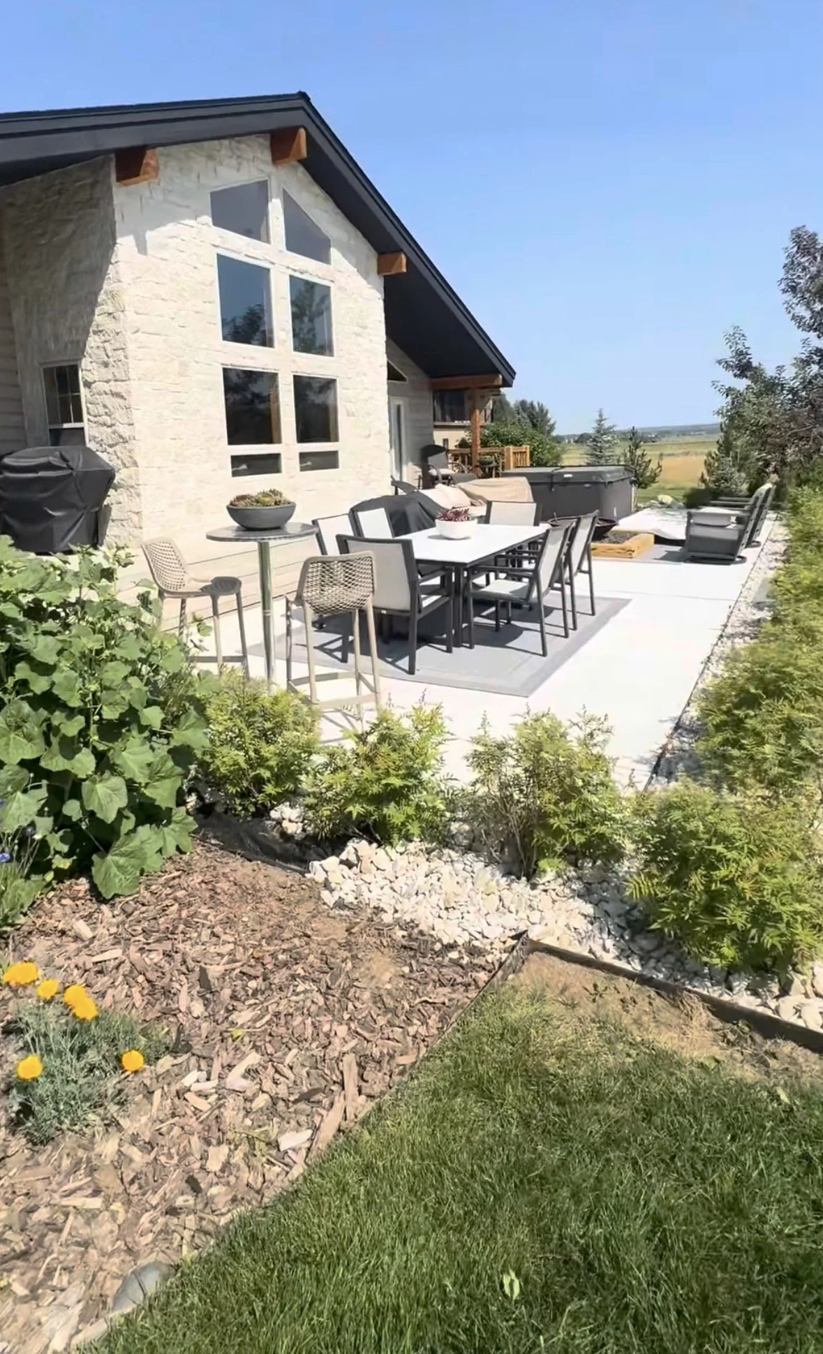 Backyard patio with outdoor dining table and chairs, garden plants, a hot tub, and a modern house with large windows and a sloped roof under clear blue sky.