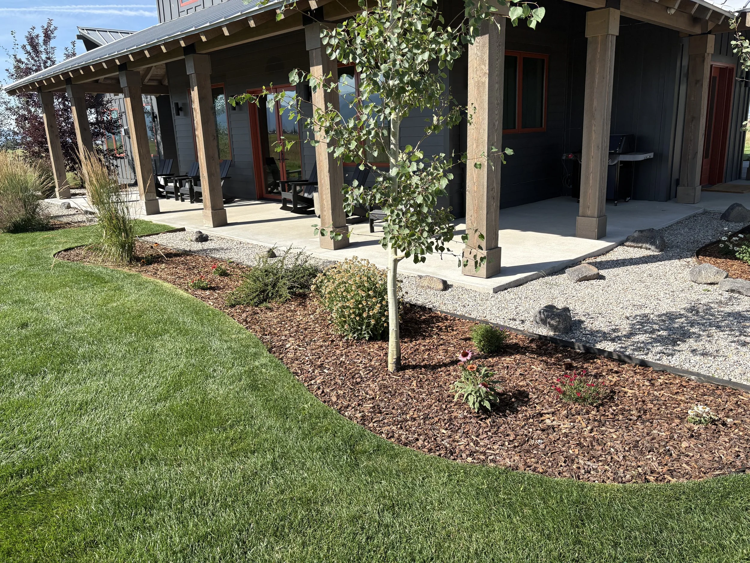 The image shows an outdoor scene with a modern house that has a covered patio area. There are several black chairs on the patio, some greenery including a small tree and various shrubs in the garden bed below. The yard has a manicured lawn, decorativ