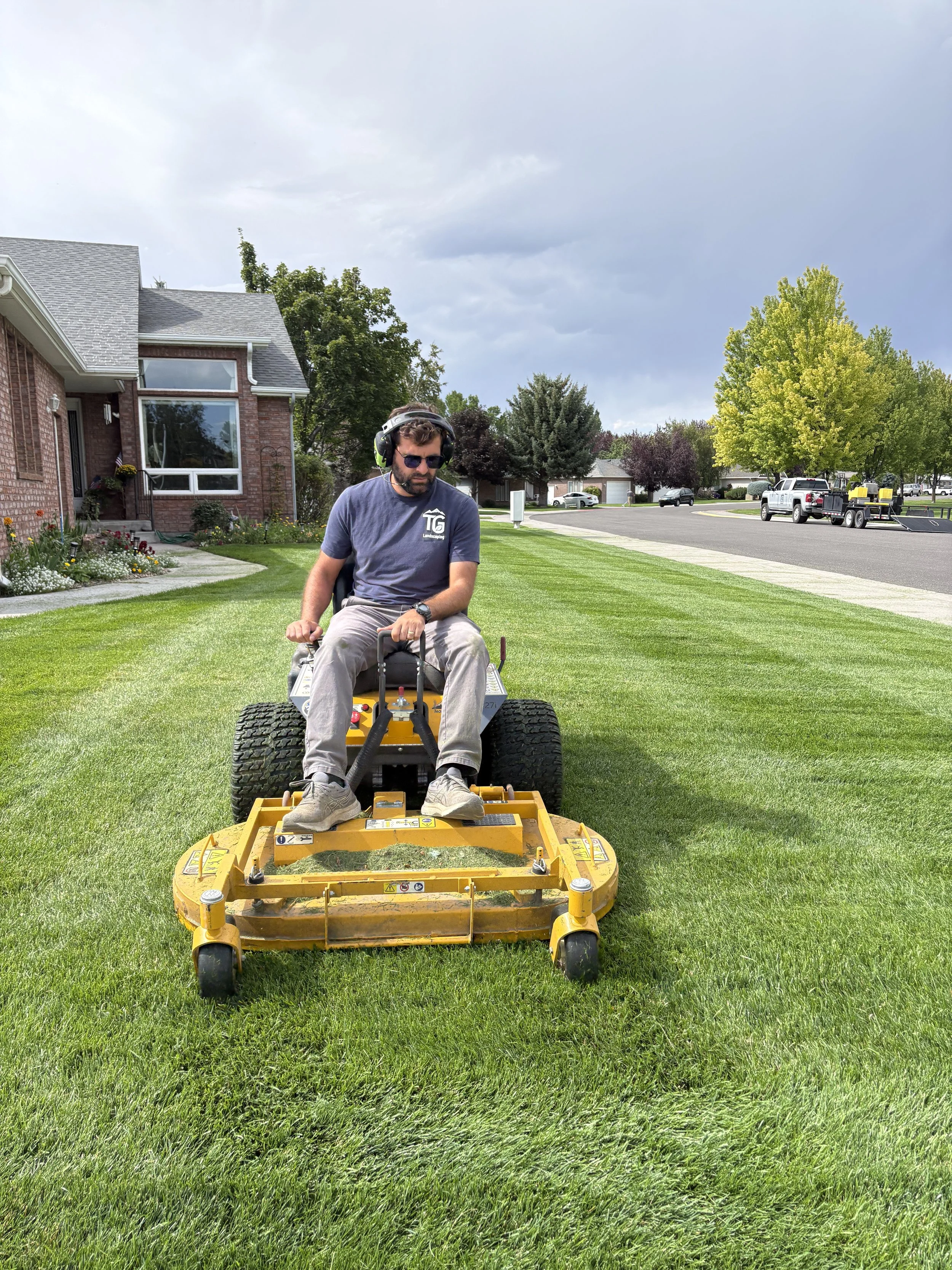 A man wearing sunglasses, a baseball cap, and headphones operates a yellow riding lawn mower on a neatly trimmed lawn in a suburban neighborhood with houses and trees under a partly cloudy sky.