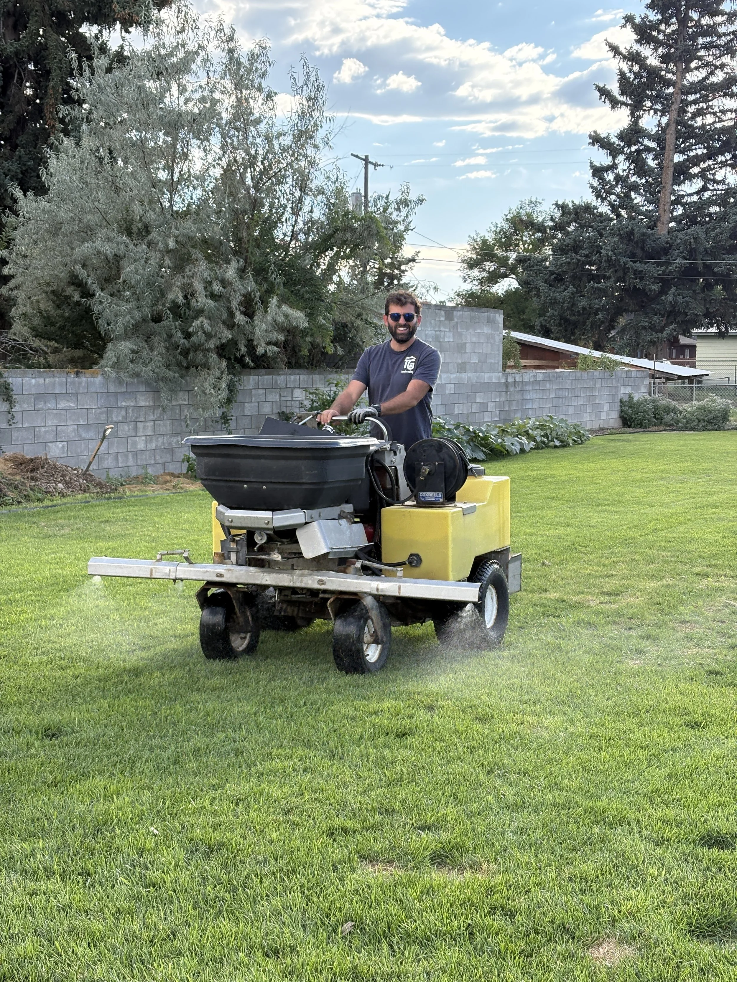 A man with sunglasses and a beard is riding a zero-turn lawn mower on a well-manicured green lawn. He is smiling and wearing a navy shirt. The background features a stone wall, trees, a blue sky with clouds, and some houses.