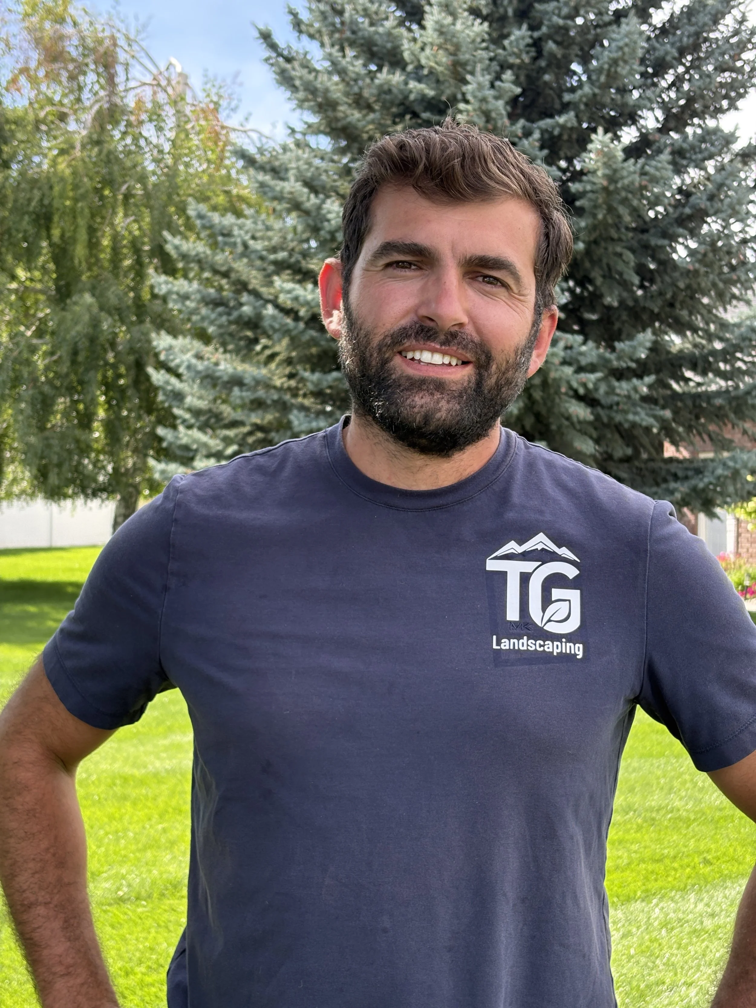 A man with dark hair and a beard wearing a navy blue T-shirt with a landscaping company logo, standing outdoors in front of a large evergreen tree, with green grass and other trees in the background.