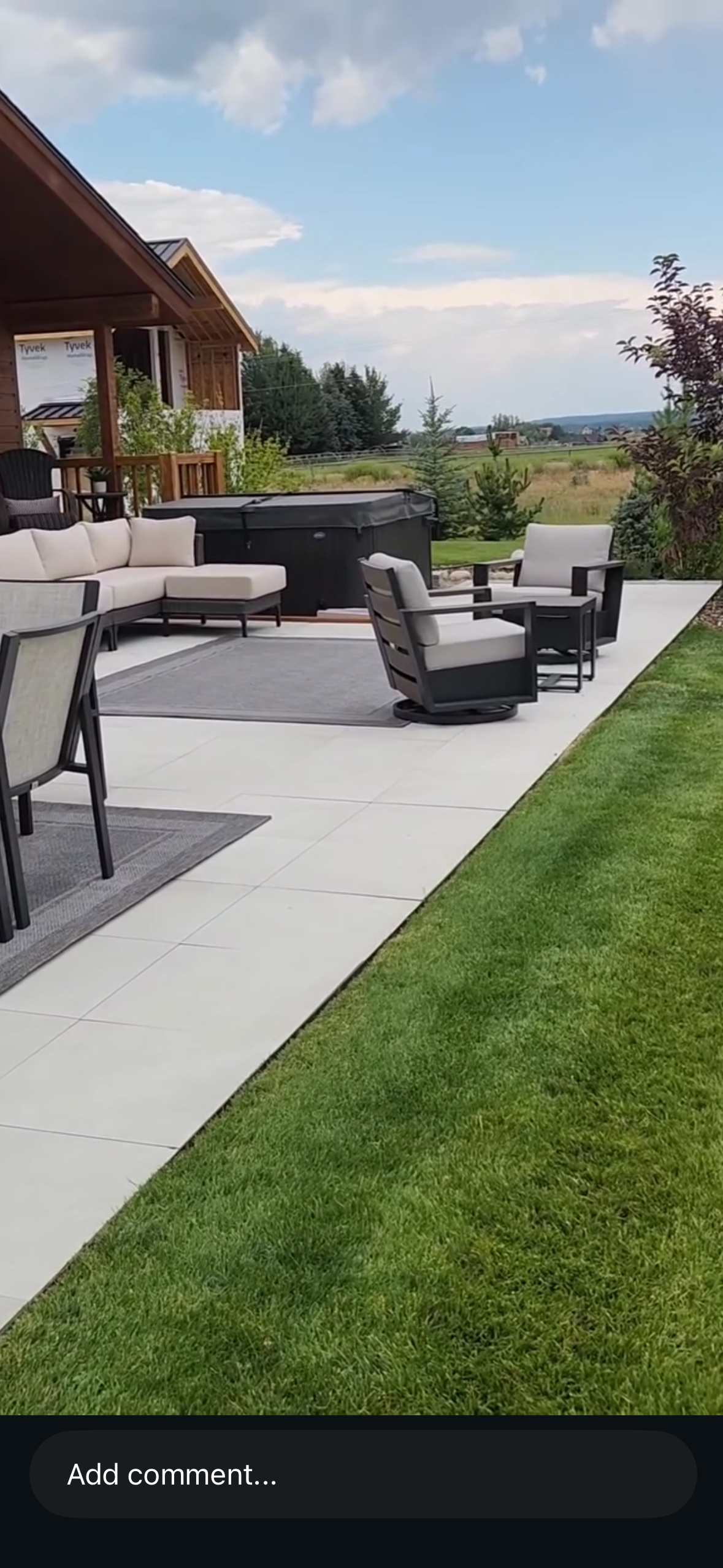 Outdoor patio with white tiled flooring, black and beige seating, a hot tub, and a grassy yard with trees and open field in the background under a cloudy sky.