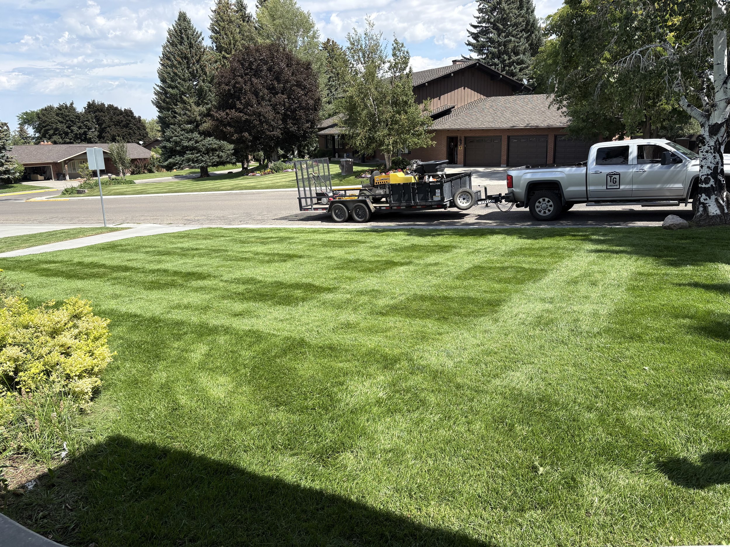 Suburban street with green lawns, trees, and houses in the background. A pickup truck towing a trailer with equipment parked on the street.