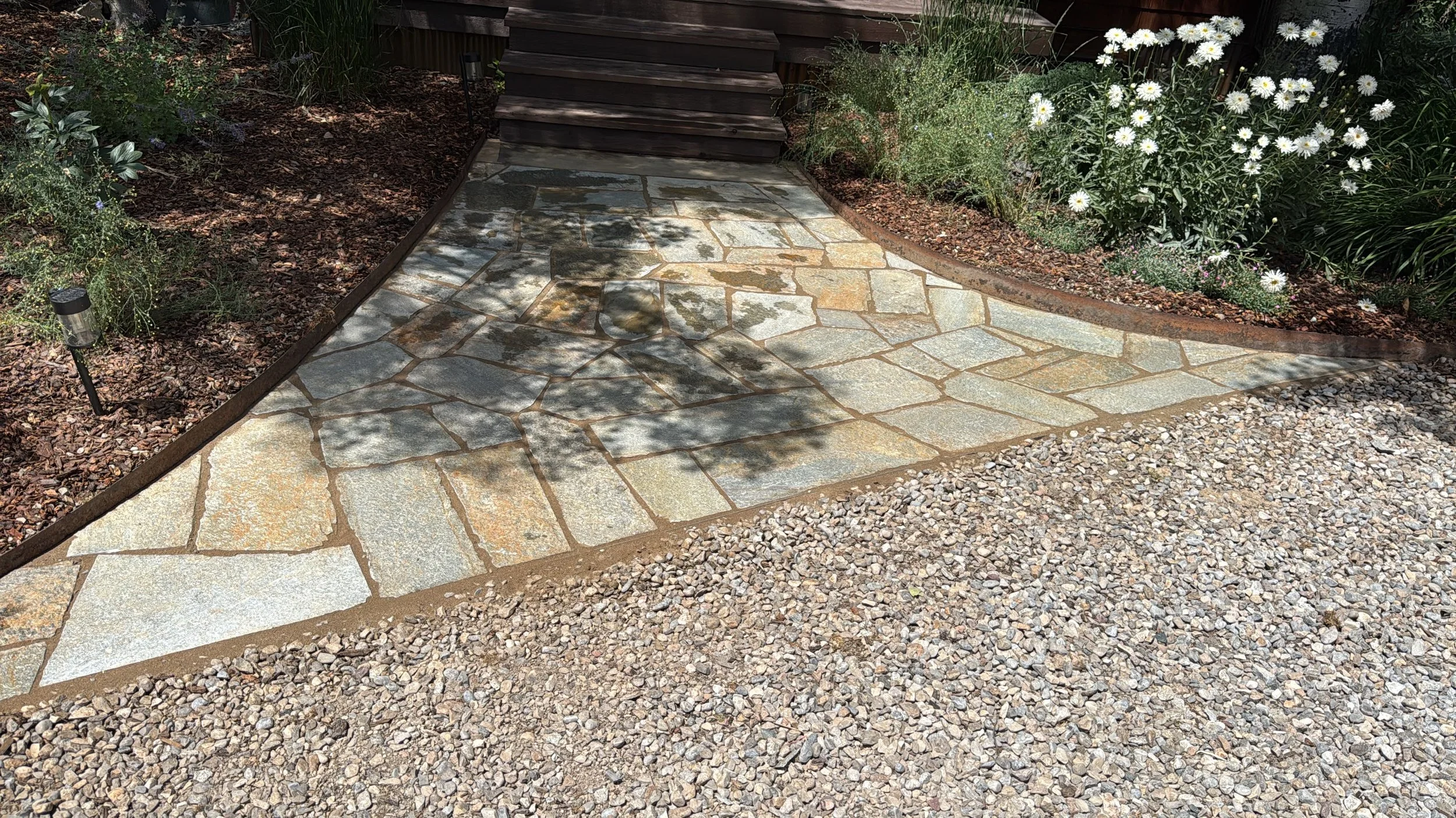 A garden pathway made of irregular-shaped paving stones leading to a staircase, surrounded by flower beds with white daisies and various green plants, with solar lights along the edges.