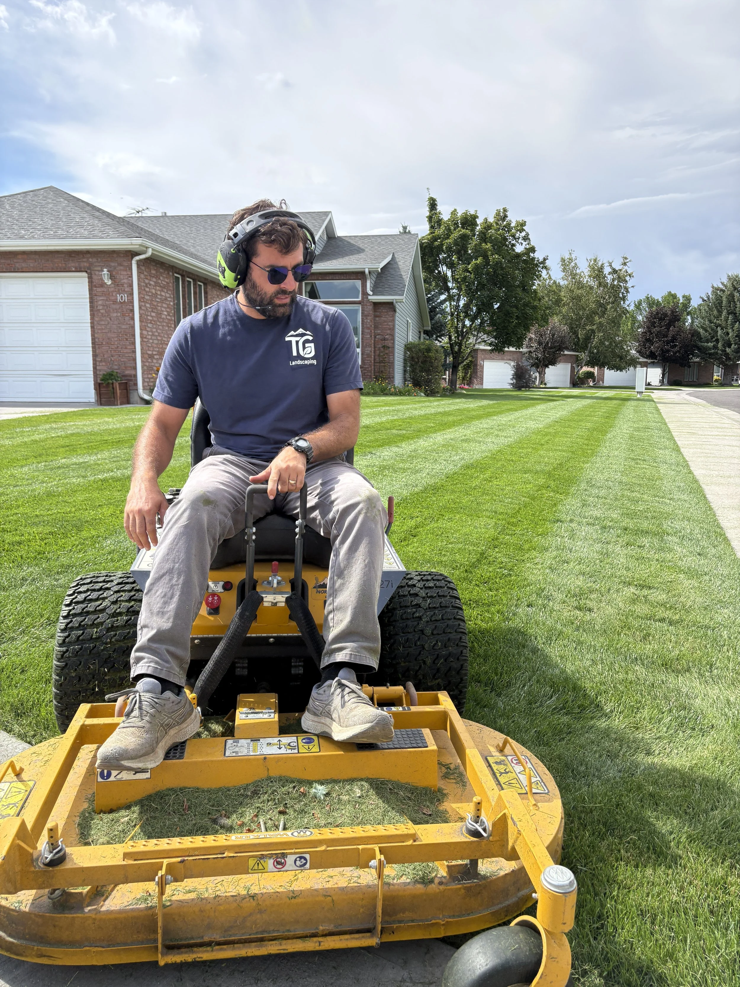A man wearing sunglasses, a headset, and a gray T-shirt sitting on a yellow lawn mower on a front yard with green grass and a sidewalk. There are houses and trees in the background under a partly cloudy sky.