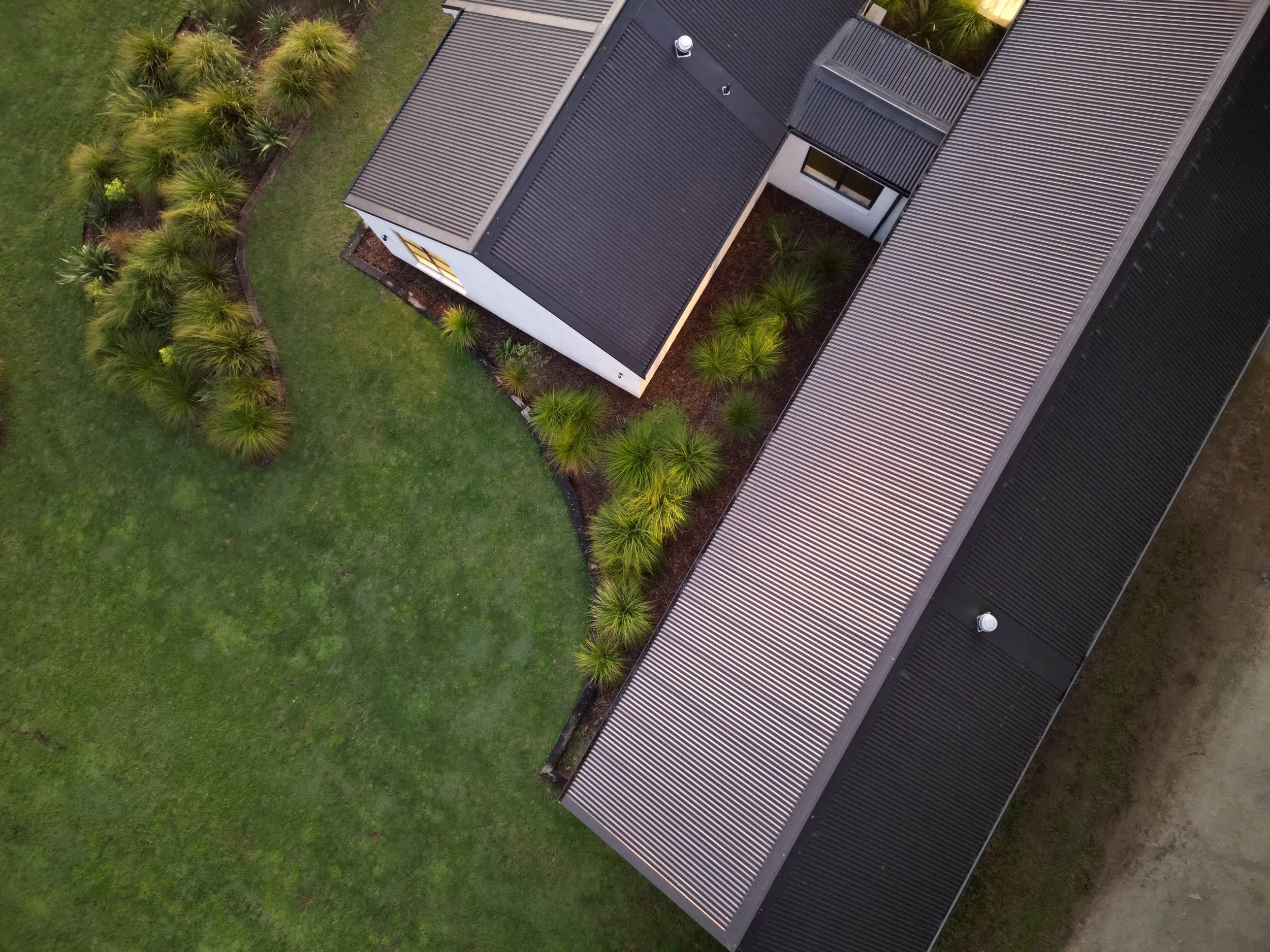 Aerial view of a modern house with a dark metal roof, surrounded by green lawn and landscaped bushes.