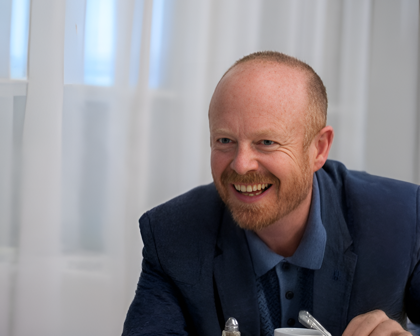 A smiling man with a beard and short hair, wearing a dark blazer and light shirt, sitting at a table.