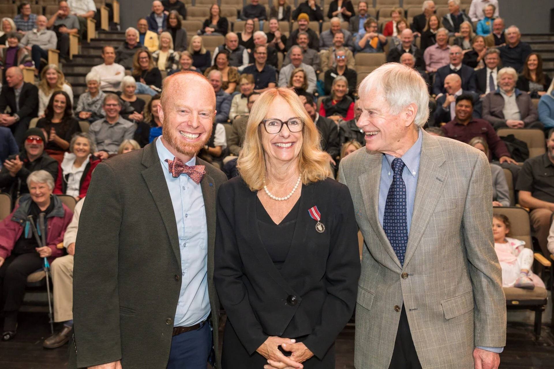 Three people standing together in front of an audience. The man on the left is wearing a dark suit with a light blue shirt and a bow tie. The woman in the middle is wearing glasses, a black outfit with a pearl necklace, and a medal pinned to her chest. The man on the right is dressed in a light gray checkered blazer, blue shirt, and patterned tie. The audience in the background is smiling and clapping, seated in an auditorium.