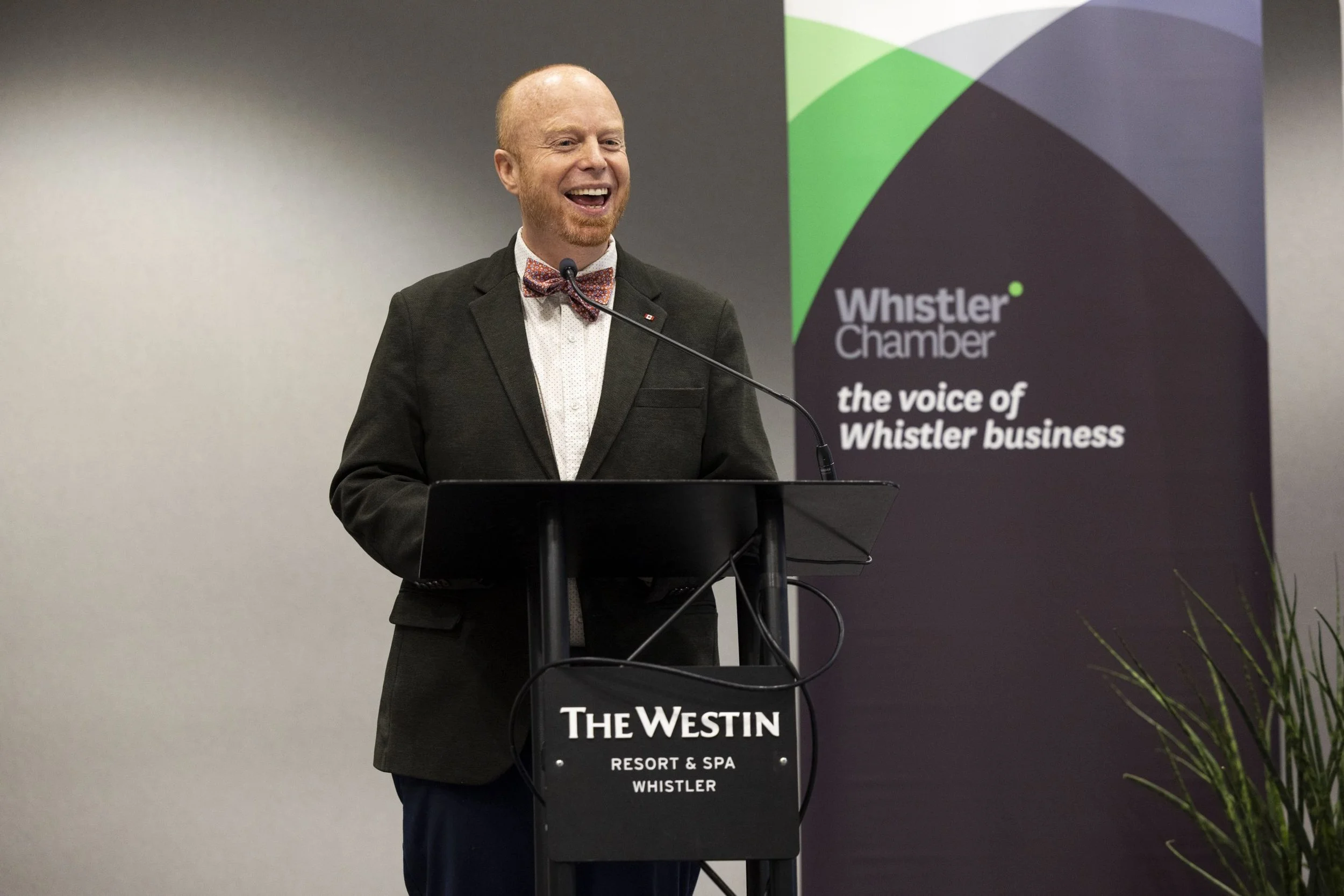 A man with a beard, wearing a suit and bow tie, speaking at a podium in front of a banner that reads 'Whistler Chamber' and 'the voice of Whistler business' at The Westin Resort & Spa in Whistler.