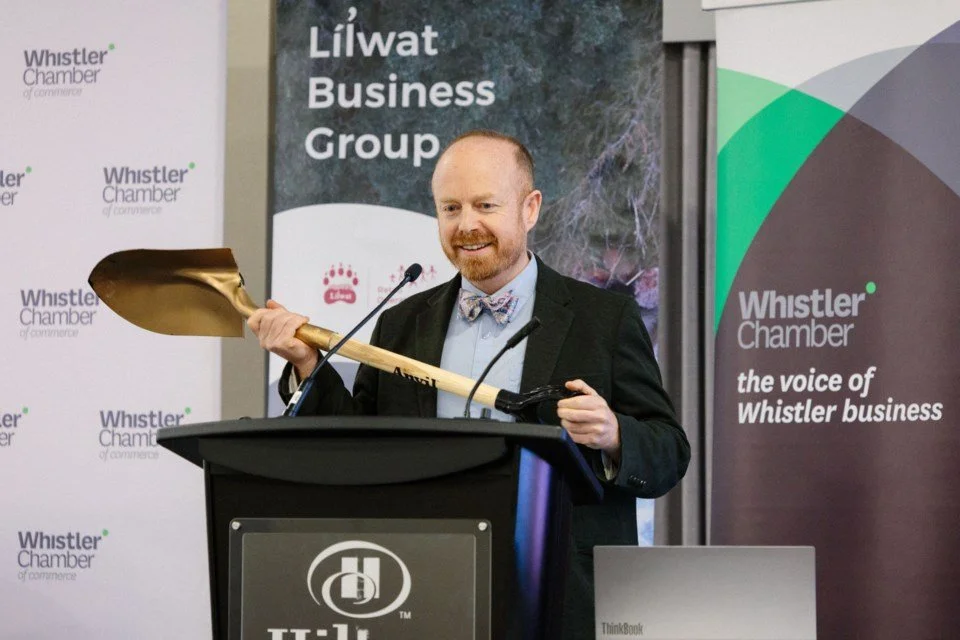 A man with a beard and red hair, dressed in a suit with a bow tie, is smiling while standing behind a podium, holding a large gold-colored shovel. Behind him are banners for the Whistler Chamber of Commerce and LiLwat Business Group, indicating a business event or ceremony.