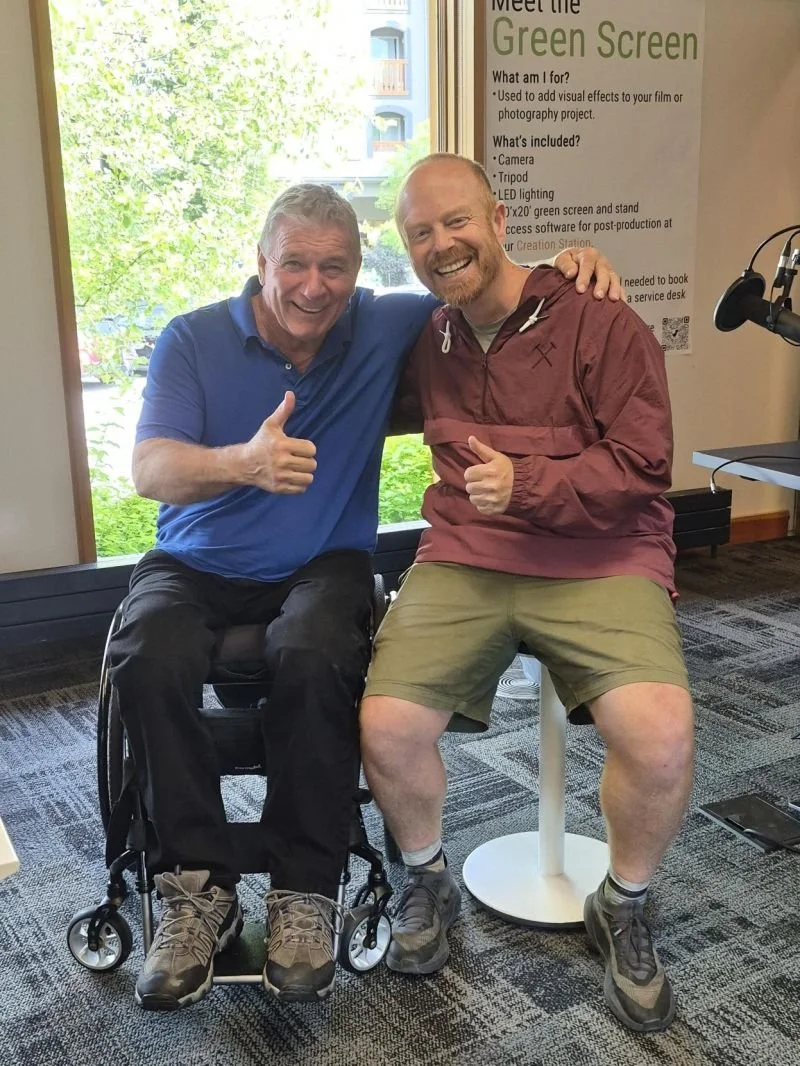 Two men sitting indoors, smiling and giving thumbs-up signs. One man is in a wheelchair, wearing a blue shirt and dark pants. The other man, wearing a red jacket and khaki shorts, is sitting on a white stool. Behind them, there's a large window with a view of green trees and a building. A poster about a green screen setup is visible on the wall.