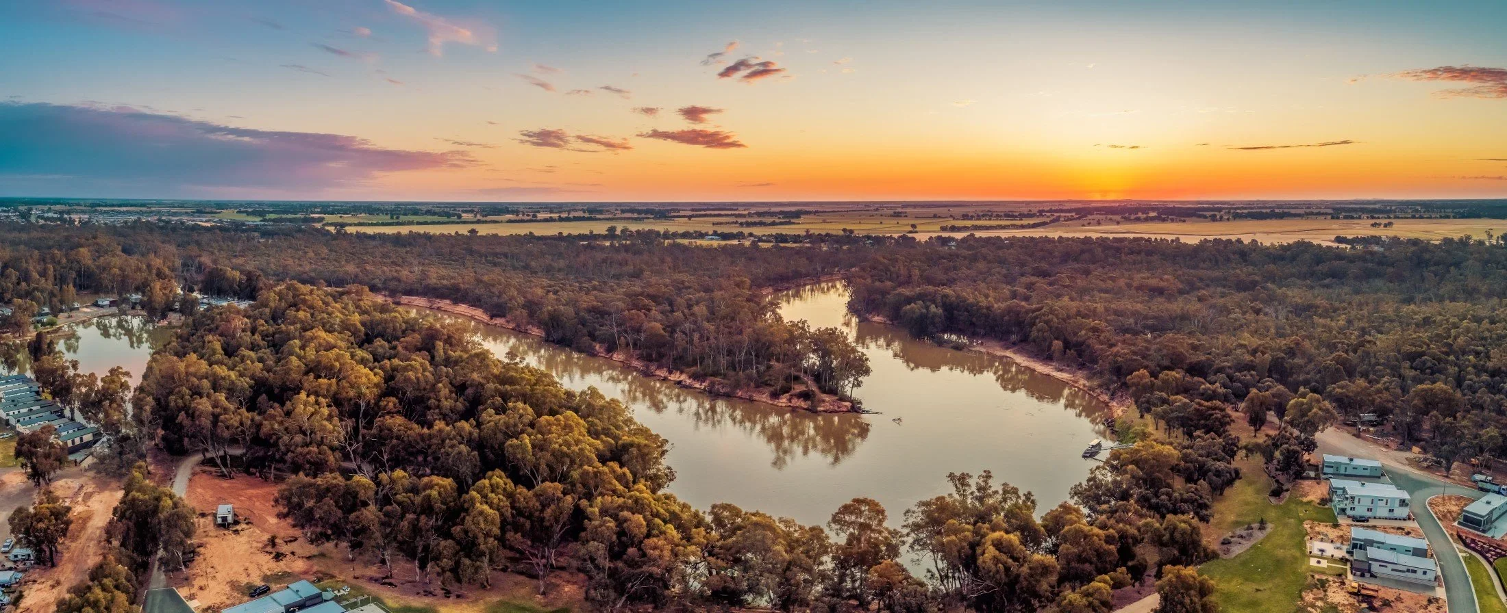 Aerial view of a winding river surrounded by trees, houses, and open fields during sunset with a colorful sky with clouds.