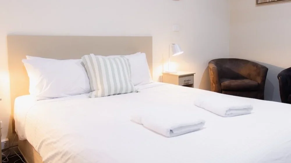 A neatly made bed with white bedding, two folded towels, and a striped pillow. There is a nightstand with a lamp and a chair in the corner of the room.