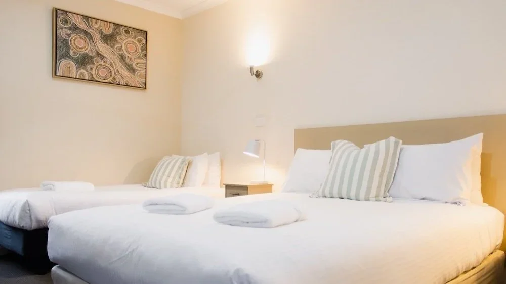Hotel room with two beds, pillows, rolled towels, nightstand, lamp, and wall art.