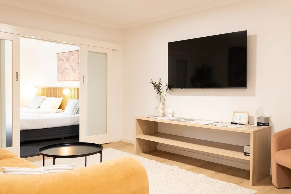 Modern living room with beige sofa, flat-screen TV on a wooden console, and a sliding door leading to a bedroom with a large bed and decorative light fixtures.