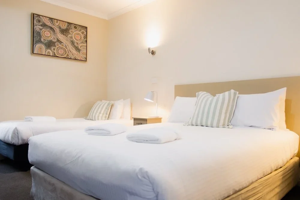 A hotel room with two beds, a nightstand, a lamp, and wall art.
