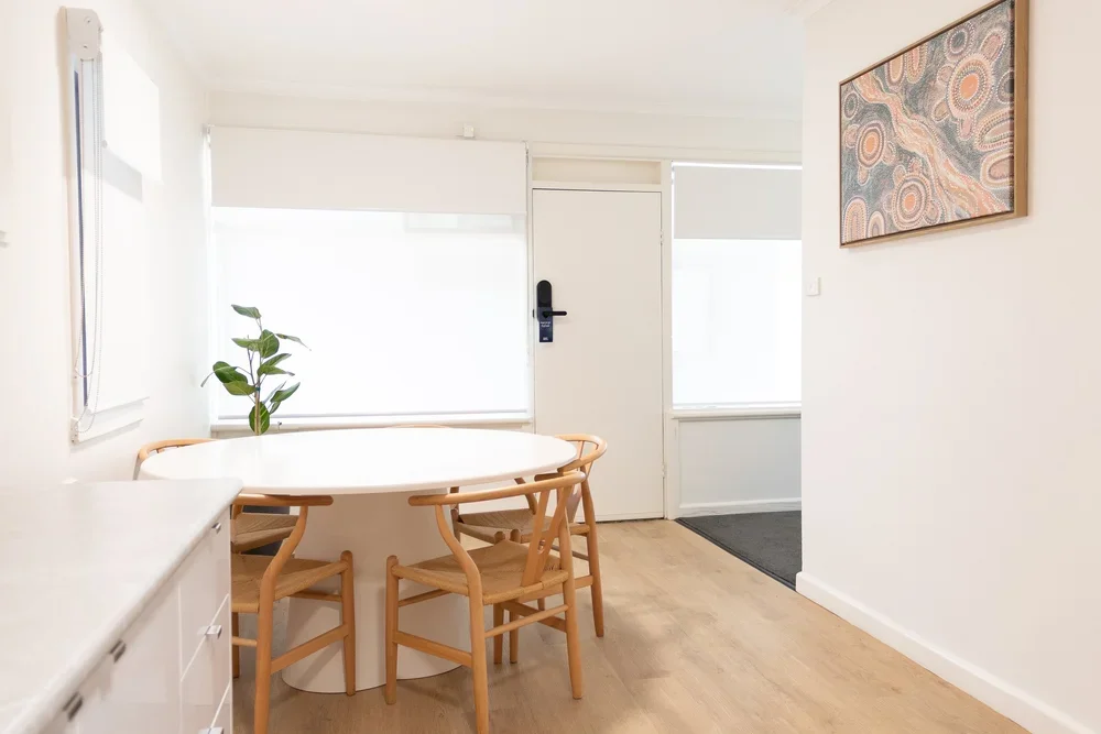 Dining room with a white oval table, four wooden chairs, a potted plant, and a large window with white blinds, next to a white door with a black handle and a colorful abstract painting on the wall.