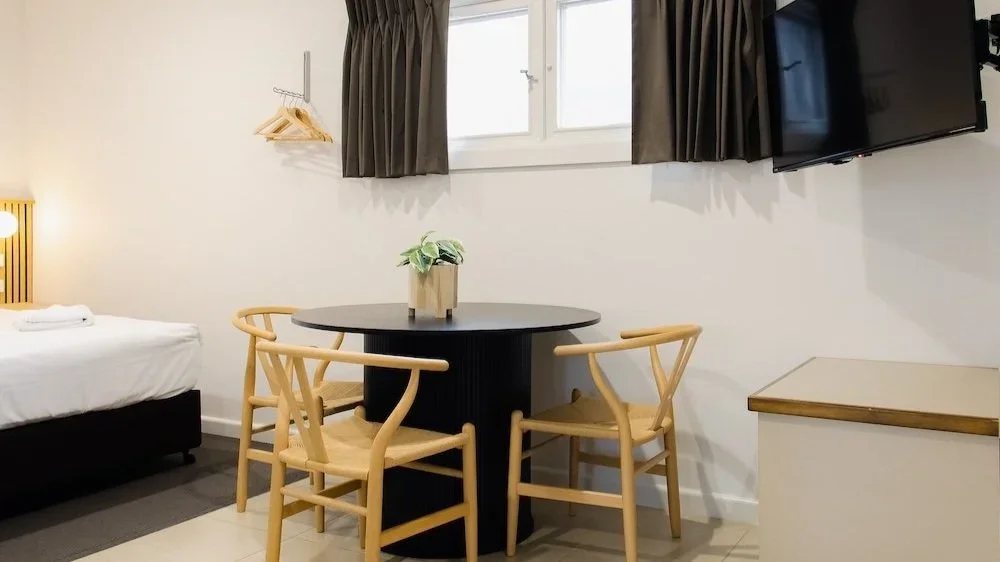 A hotel room with a bed, a small round dining table with four chairs, a wall-mounted TV, and a window with curtains.