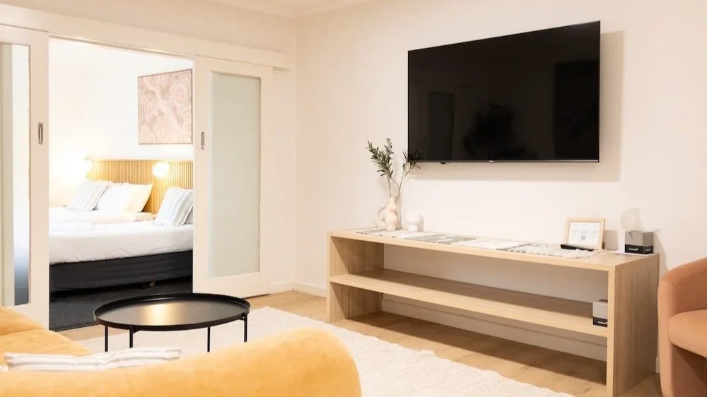 Living room with a wall-mounted TV, a light wood TV stand, a vase with greenery, and a large sliding glass door leading to a bedroom with a bed and nightstand.