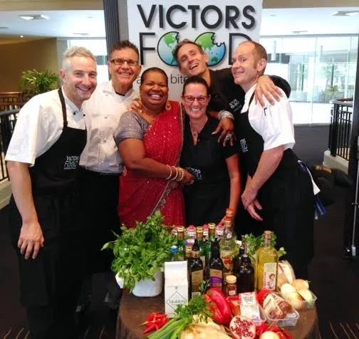 Group of six people in a restaurant or cafe, smiling and posing together, with a table of food and drinks in front of them. A sign in the background reads 'VICTORS FOOD'.