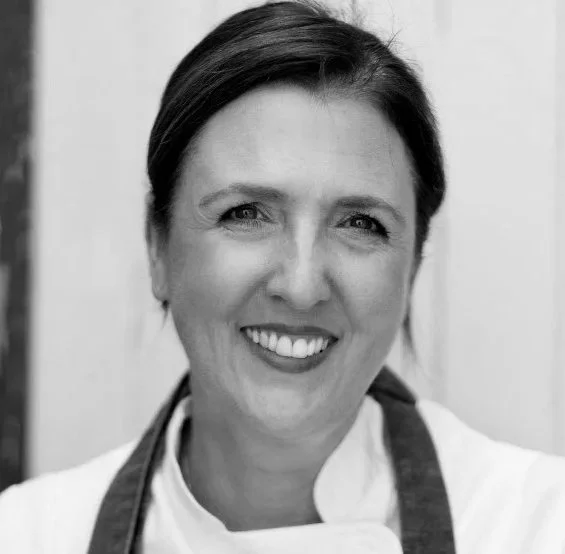 Close-up black and white portrait of a smiling woman with short dark hair, wearing a white chef's jacket and apron.