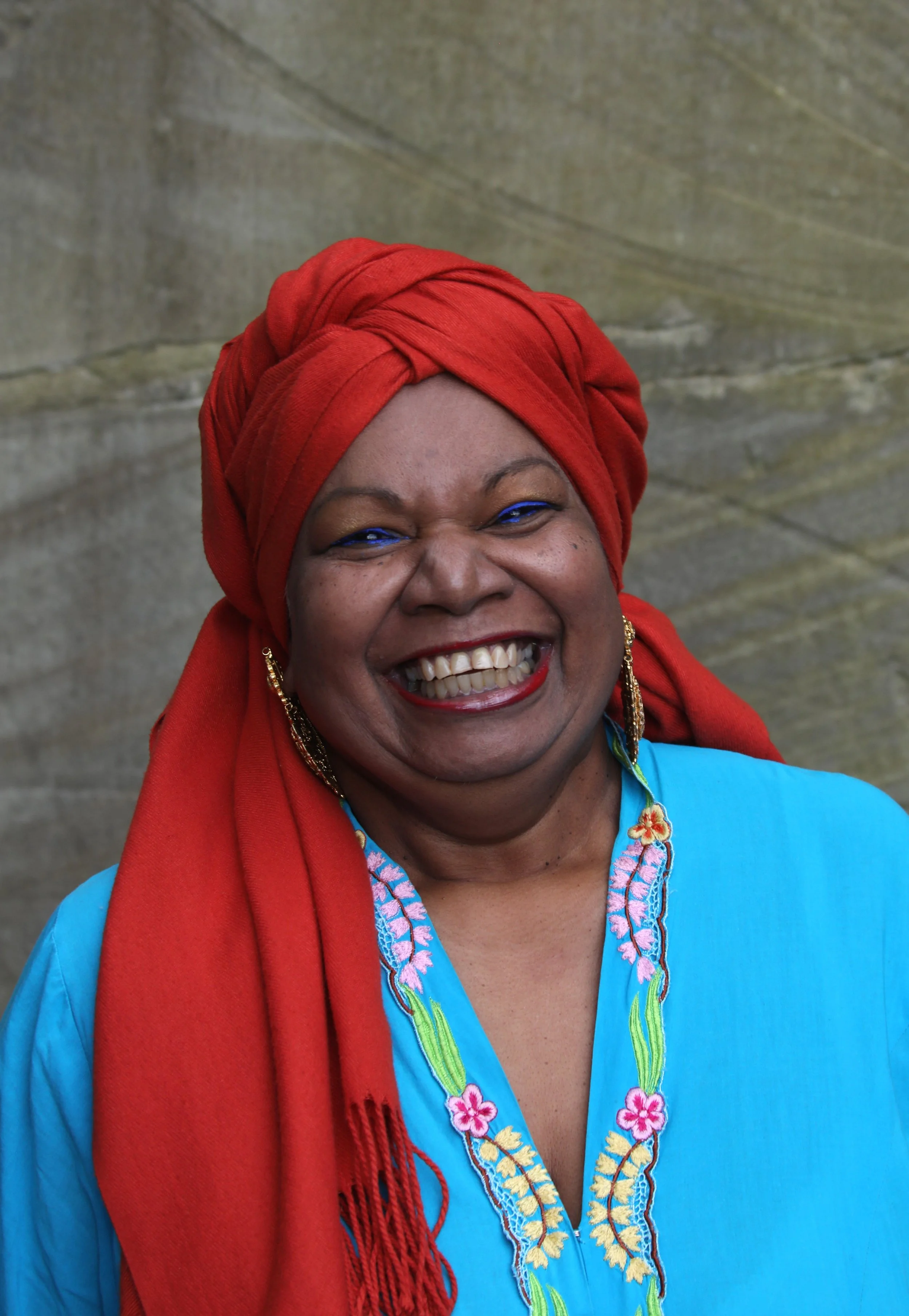 A smiling woman wearing a bright red head wrap and a blue embroidered top, standing against a stone wall.