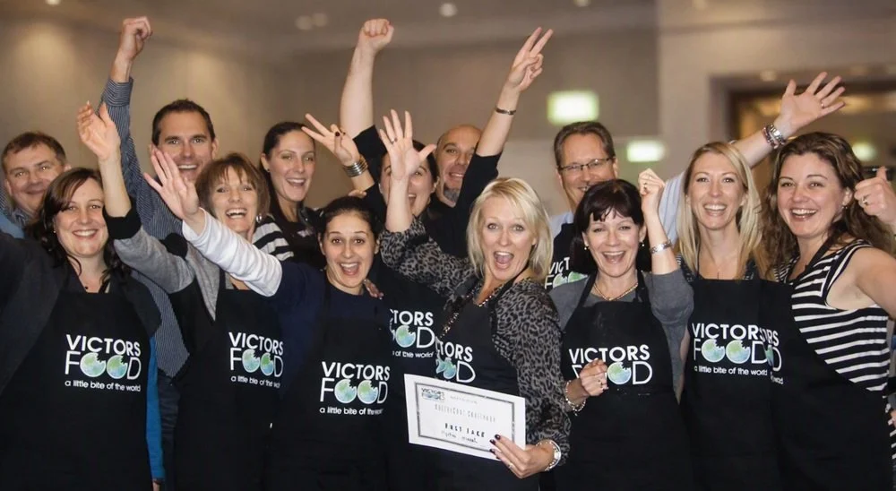 Group of smiling people wearing chefs aprons celebrating a team building cooking challenge, with one person holding a certificate, in an indoor setting.